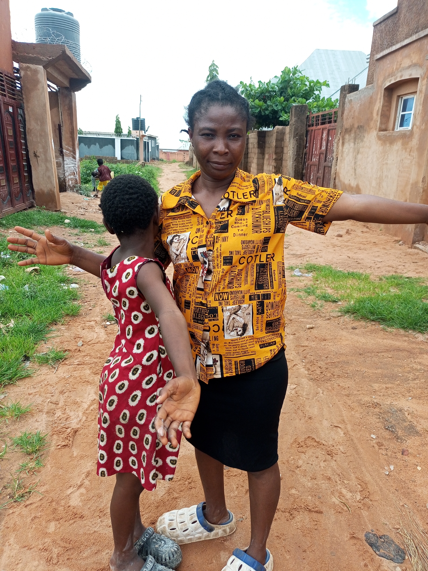 In Tempure, Nigeria, a woman and a young girl stand on a dirt road. The woman, with dark hair pulled back, wears a yellow short-sleeved shirt with a newspaper print and a black skirt. She has her arms outstretched to her sides. The girl, facing away from the camera, has dark, curly hair and wears a red sleeveless dress with a white and black pattern. Her left arm is extended, and her right arm is bent. Both are wearing sandals. The background shows a partially built structure, a large water tank on a platform, and some vegetation.
