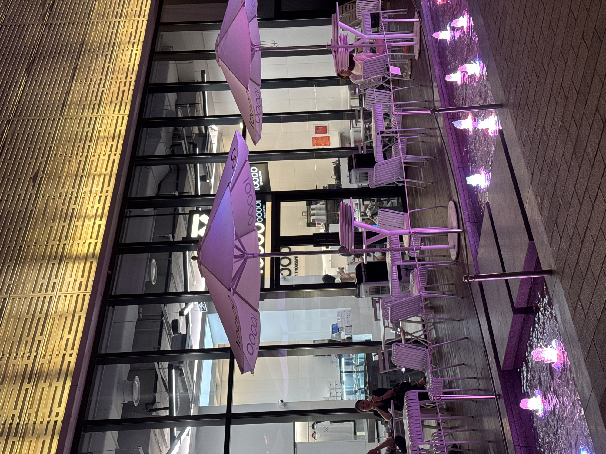 This is a nighttime photograph of an outdoor seating area for a restaurant or cafe. The seating area features numerous white metal tables and chairs arranged on a wooden deck. Two large purple umbrellas provide shade. In the foreground, a water feature with illuminated fountains adds a decorative element. Behind the seating area, a modern building with large glass windows is visible. The building's facade is adorned with a slatted design illuminated by yellow lights. Several people are seated at the tables, some interacting with each other, while others appear to be eating or drinking. The scene is illuminated by artificial lighting, casting purple and yellow hues. The presence of a water feature and outdoor seating suggests a public or commercial establishment in a city. Based on the architectural style and the presence of the brand "10000 FOOD AND THOUSAND," this is likely a cafe or restaurant in Hanoi, Vietnam, possibly in a modern commercial district.