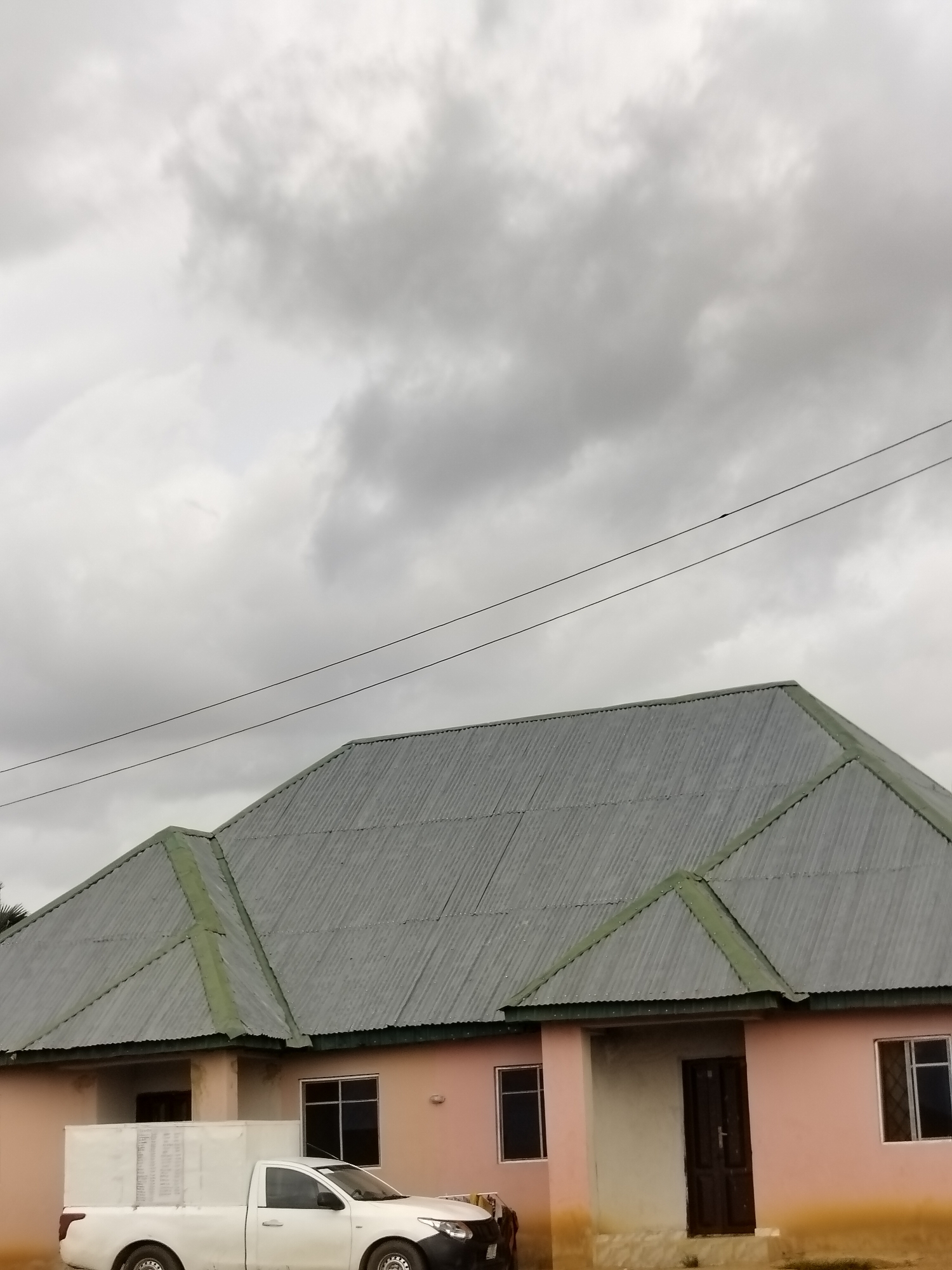 The image depicts a light pink building with a corrugated metal roof, situated in Du, Nigeria. The building has several windows and a dark wooden door. In front of the building is a white pickup truck with a large white rectangular box mounted on its bed. The sky above is cloudy and gray. The scene appears to be an outdoor view of a residential or commercial property.