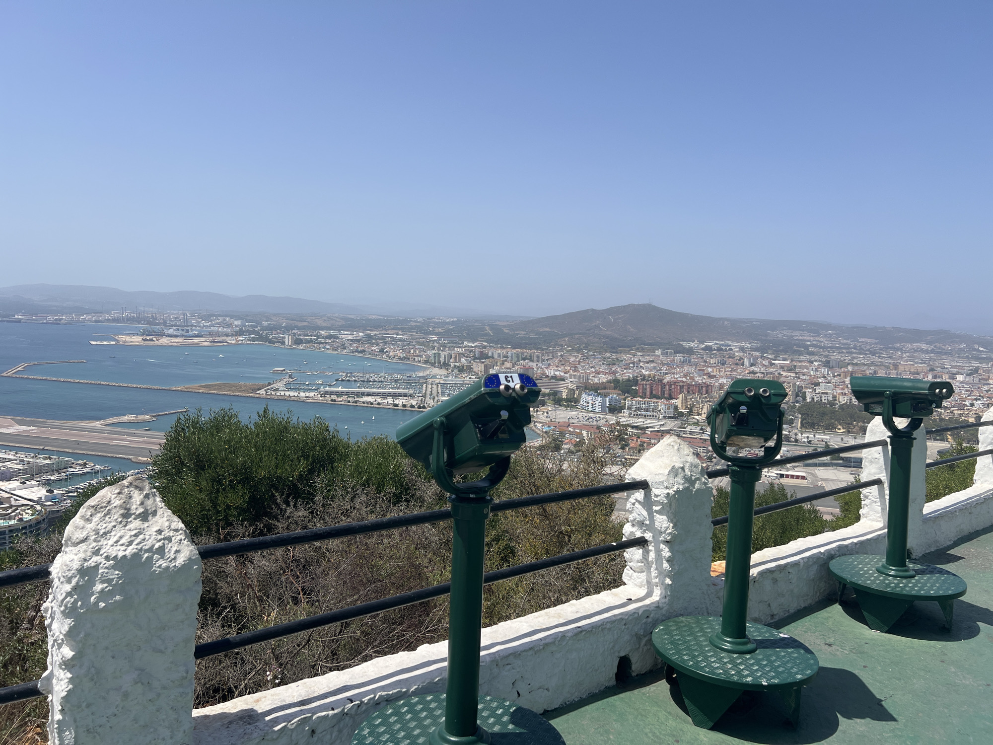 The image captures a panoramic view from a high vantage point in Gibraltar. In the foreground, three green coin-operated binoculars stand on a concrete platform, facing the expansive vista. The binoculars are arranged in a row, with the leftmost one being the closest to the viewer. They are mounted on sturdy green pedestals with textured circular bases. A white textured wall with a black metal railing runs along the edge of the viewpoint, separating it from the drop below.
Beyond the railing, a lush green tree sits to the left, with dry, thorny bushes to its right. The city of Gibraltar stretches out below, a dense urban landscape with numerous buildings of various heights and colors, predominantly white and orange, clustered together. A harbor is visible on the left, filled with boats and yachts, with an airport runway extending into the sea. Further in the distance, a bay connects to the open sea, and beyond that, hazy hills and mountains form the backdrop under a clear blue sky. The overall scene depicts a tourist viewpoint offering a sweeping outlook of the city, harbor, and surrounding natural landscape.