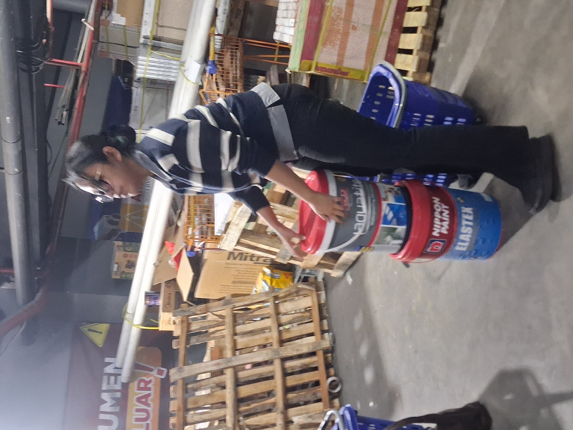 In Depok, Indonesia, a young woman wearing a blue and white striped sweater, black pants, and glasses stands in what appears to be a warehouse or large retail store. She is holding a stack of three paint buckets. The top bucket is gray with white lettering, the middle is red with white lettering, and the bottom is blue with white lettering. To the left of the woman is a stack of wooden pallets and a blue plastic basket. In the background, there are more pallets, shelves, and what seem to be large pipes. A sign with yellow lettering and a yellow warning triangle is visible in the far background.