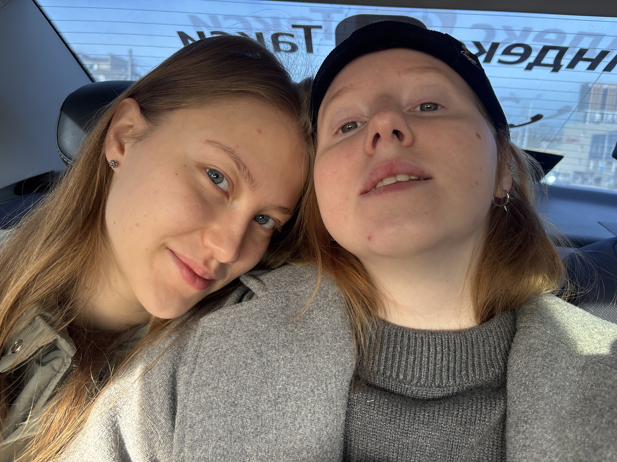 Two young women are posing for a selfie inside a car. The woman on the left has long, light brown hair and blue eyes. She is wearing a light-colored jacket. The woman on the right has reddish-blonde hair and is wearing a black beanie and a grey sweater underneath a grey coat. The background visible through the car window suggests they are in Perm, Russia, indicated by the Cyrillic lettering "ЯНДЕКС ТАКСИ" (Yandex Taxi) on the rear window. The vehicle's interior is visible with the headrest and seat.