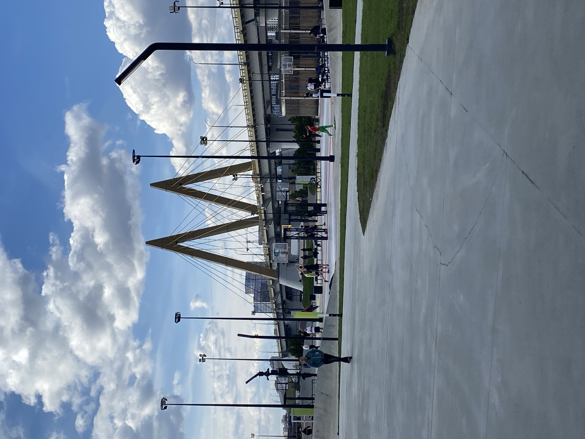 The image displays an outdoor urban scene in Kazan, Russia, under a bright, partly cloudy blue sky. The most prominent feature is the Millennium Bridge in the background, recognizable by its two large, A-shaped pylons of a golden or light yellow hue, from which numerous cables descend to support the bridge deck. Below the main bridge structure, further urban infrastructure and buildings are visible.
In the foreground and midground, a wide, light gray concrete pathway or plaza stretches across the frame, with a strip of green grass running alongside it. Numerous tall, black lampposts, some with ring-shaped lights and others with rectangular floodlights, are positioned along the path and grass areas.
Several people are engaged in various activities within this public space. One individual in a red shirt and green pants is captured in mid-air or mid-stride on a reddish court area, suggesting athletic activity or play. Another person wearing a dark teal/green top stands further down the concrete path. Other figures are discernible near what appears to be outdoor fitness equipment and a basketball hoop. A bicycle is also parked in the midground.
The time of day is clearly daytime, marked by the brilliant sunlight and the presence of fluffy white cumulus clouds against the clear blue sky, indicating fair weather. The scene conveys an impression of a modern, active recreational area adjacent to a significant architectural landmark. No visible text is legible in the image.