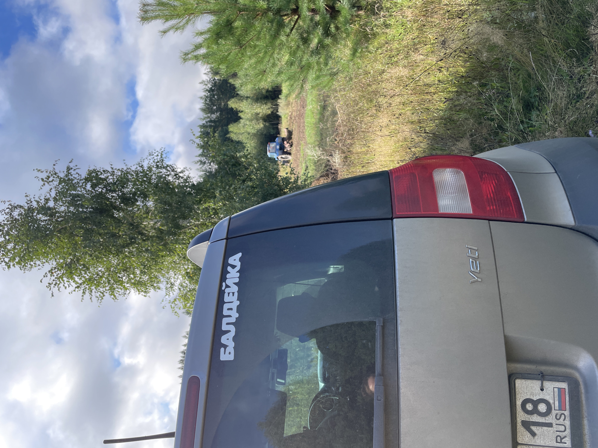 The image shows the rear of a silver Skoda Yeti parked on a grassy area with dry grass and some trees. A blue tractor is visible in the background, plowing a field. There is a white sticker on the back window of the car that reads "БАЛДЕЙКА". The license plate of the car shows "18 RUS". The sky is partly cloudy. The scene appears to be in a rural area in Russia, with the text on the sticker indicating "Baldieyka" as a location.