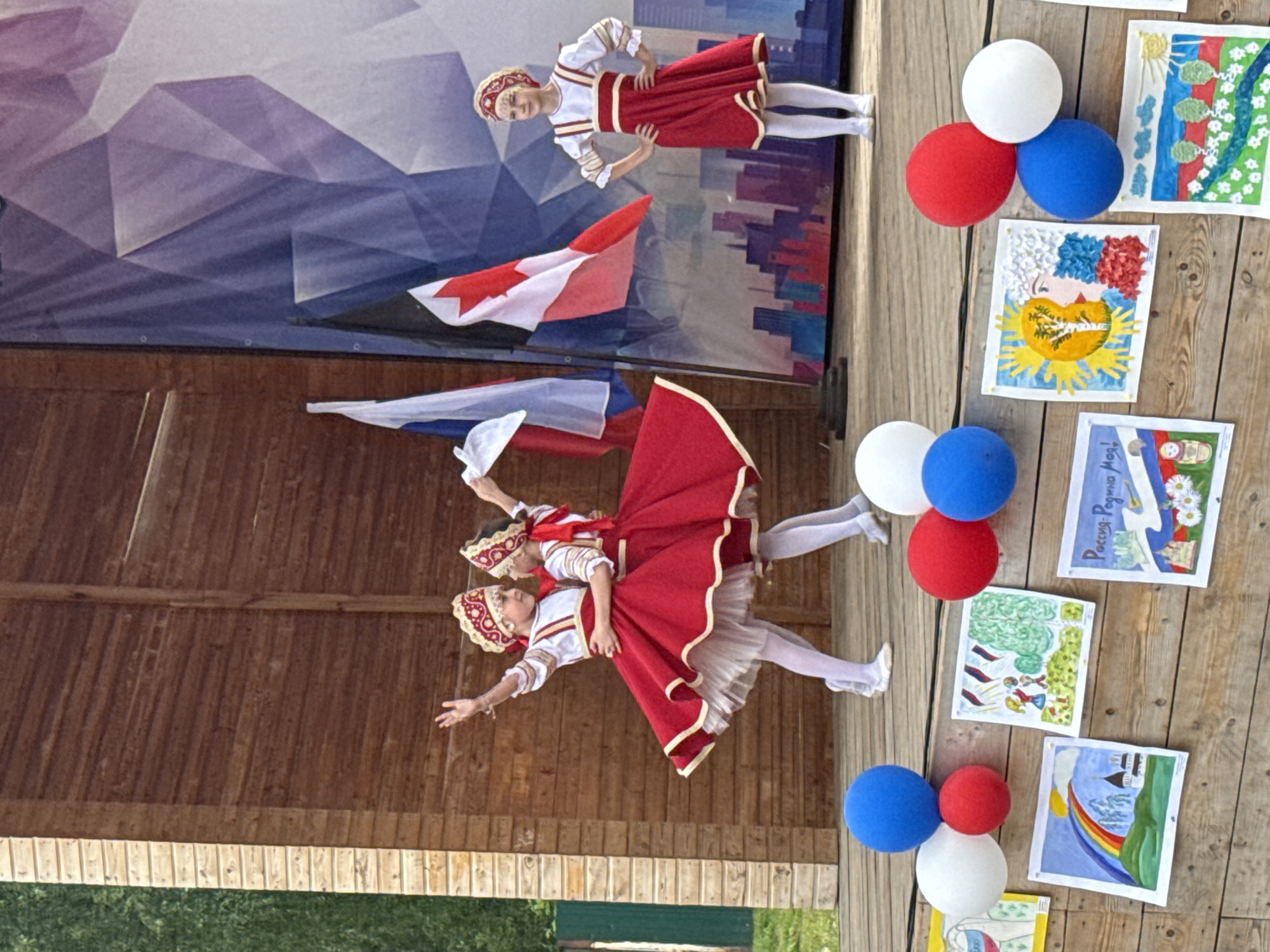 On a wooden stage, three young girls in traditional Russian folk costumes are performing. Two girls in the center, adorned in red dresses with white and gold trim and matching headwear, are dancing together, their arms extended. A third girl, standing to the right in a similar red and white outfit with a headband, poses with her hands on her hips. Behind them, flags of Russia and Canada are displayed against a backdrop depicting a stylized cityscape. The stage is decorated with colorful drawings and balloons. This event appears to be taking place outdoors in Votkinsk, Russia, possibly during a cultural festival or performance.