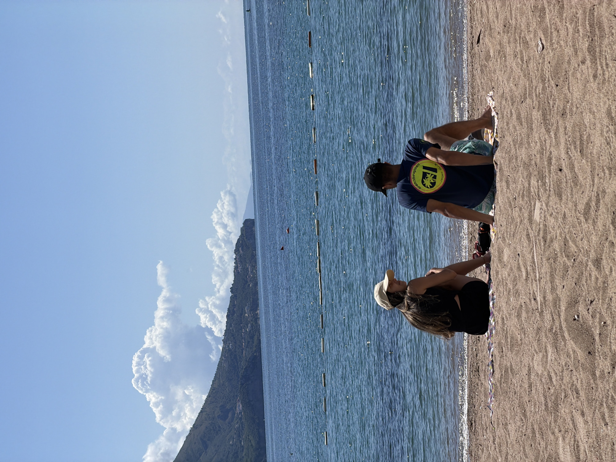Two people are sitting at the shoreline of a beach on a sunny day. A man in a navy blue t-shirt, patterned shorts, and a black baseball cap is seated cross-legged on a patterned towel. Next to him is a woman with long brown hair, wearing a black top and shorts and a tan baseball cap. She also sits cross-legged. Between them and the water is a strip of thin, multicolored ribbon or streamers.
The beach is light brown sand. The sea is a deep blue. Behind the beach and sea is a green, mountainous landmass with a clouded sky behind it. The sky is a clear, light blue.
Based on the location's geography, this scene likely takes place on one of the beaches around the city of Budva, Montenegro, given the mountainous backdrop and the coastal setting.