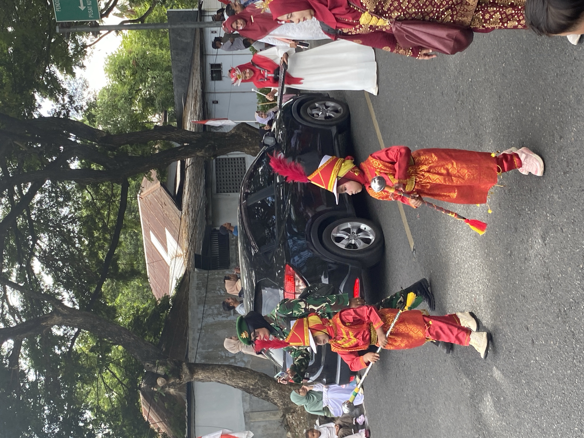 In Banda Aceh, Indonesia, a street parade is underway. The foreground features two young children in traditional red and gold marching band attire, complete with feathered hats. One child holds a drumstick. Behind them, a woman in a military uniform, wearing a green beret, takes a photo with her phone. A black SUV is parked nearby. Further back, several women in red and white traditional clothing, some wearing headscarves, are also part of the parade. The street is lined with buildings, trees, and a traffic sign pointing to "PUSAT KOTA" (City Center). The overall scene depicts a festive cultural event taking place in an urban setting.