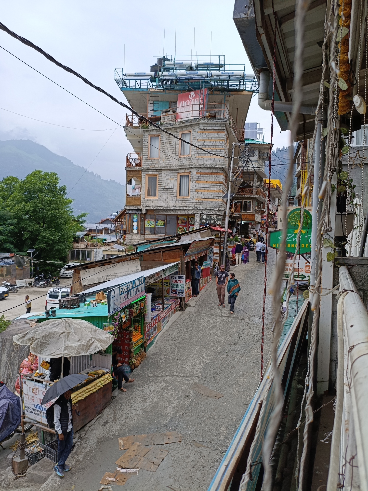 This is a vertical, eye-level, medium shot capturing a narrow street scene in Vashisht, India. The street slopes downwards from right to left and is lined with multi-story buildings and shops. A cloudy, overcast sky hangs above, with misty mountains visible in the background. Electrical wires crisscross overhead, adding to the cluttered urban feel.
On the right side of the street, a tall, stone-faced building dominates the view. It has multiple balconies and windows, and appears to be a hotel or guesthouse, indicated by a sign that reads "HOTEL VALLEY OF GODS." Further down, a shop with a green awning displays a large sign with a green tree logo and the word "Nirvana." Other shops on this side sell various goods, with awnings and displays of food items like fruits and corn.
On the left side of the street, a street vendor is set up under a large white umbrella. They are selling corn on the cob from a cart, and a person holding a black umbrella stands nearby. Further down the street, more shops and buildings are visible, with some motorcycles and cars parked.
Several people are walking along the street. Two individuals, a man and a woman, are walking down the street in the mid-ground. Another person, a man, is seen walking on the left side of the street near the vendor. In the distance, a small group of people are seen walking uphill towards the taller buildings.
The overall impression is of a bustling, somewhat chaotic, but vibrant street in a mountain town, likely a popular tourist destination. The architecture suggests a blend of traditional and modern styles, with many buildings constructed with local stone. The presence of shops, food vendors, and hotels indicates economic activity catering to visitors.