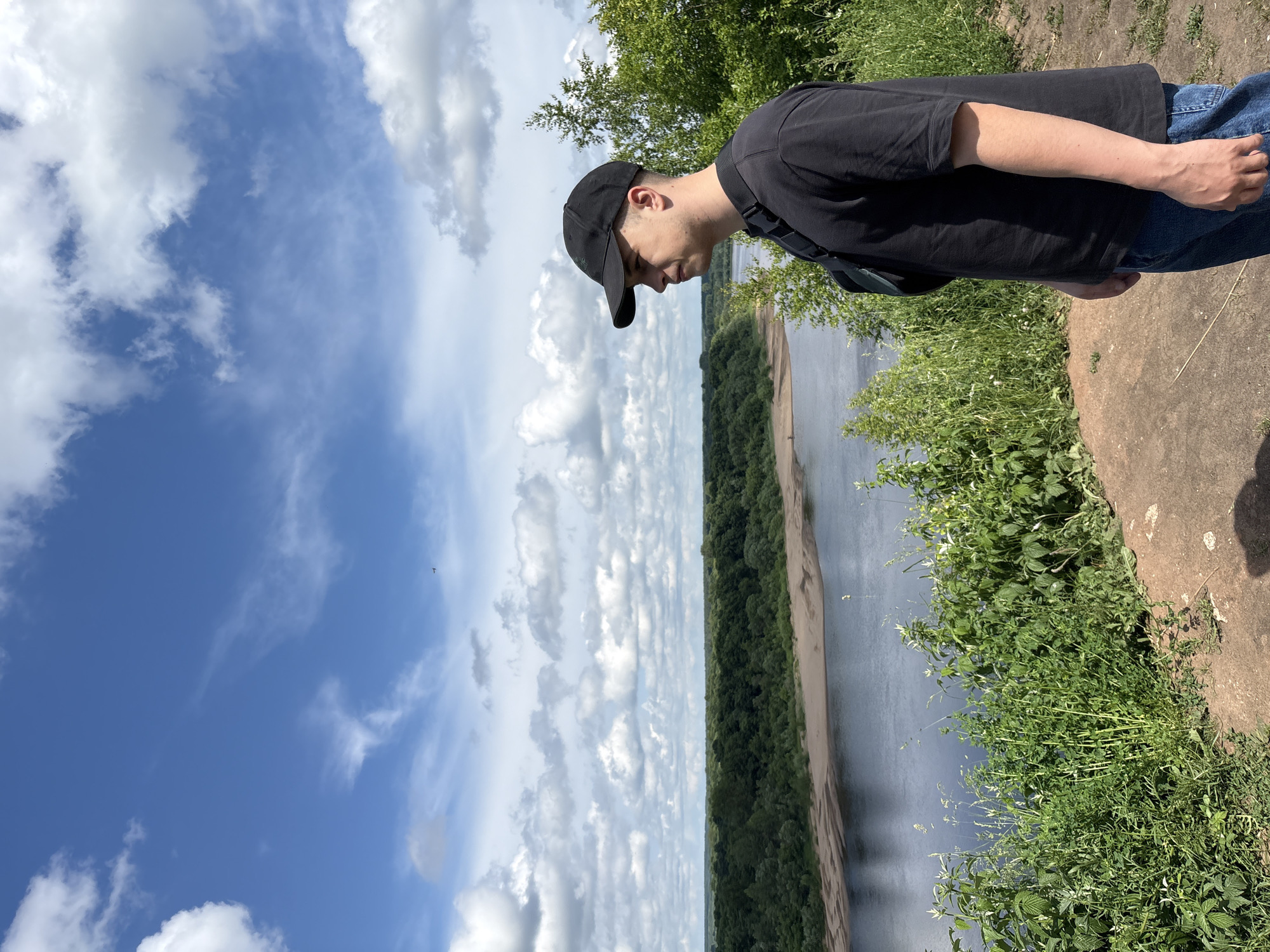 A young man wearing a black t-shirt and a black baseball cap is standing on a grassy embankment overlooking a wide river. He is looking down and to his right, with a pensive or contemplative expression. The river is bordered by a sandy bank on the opposite side, which is covered in dense green trees. The sky above is a vibrant blue, dotted with fluffy white clouds. The sun appears to be high in the sky, suggesting it is daytime. The overall setting is natural and serene, with lush vegetation and a calm body of water. The man is casually dressed, and he has a backpack slung over his shoulder. The image conveys a sense of peace and quiet observation of the natural landscape. There is no visible text in the image. The location is Kirovo-Chepetsk, Russia.