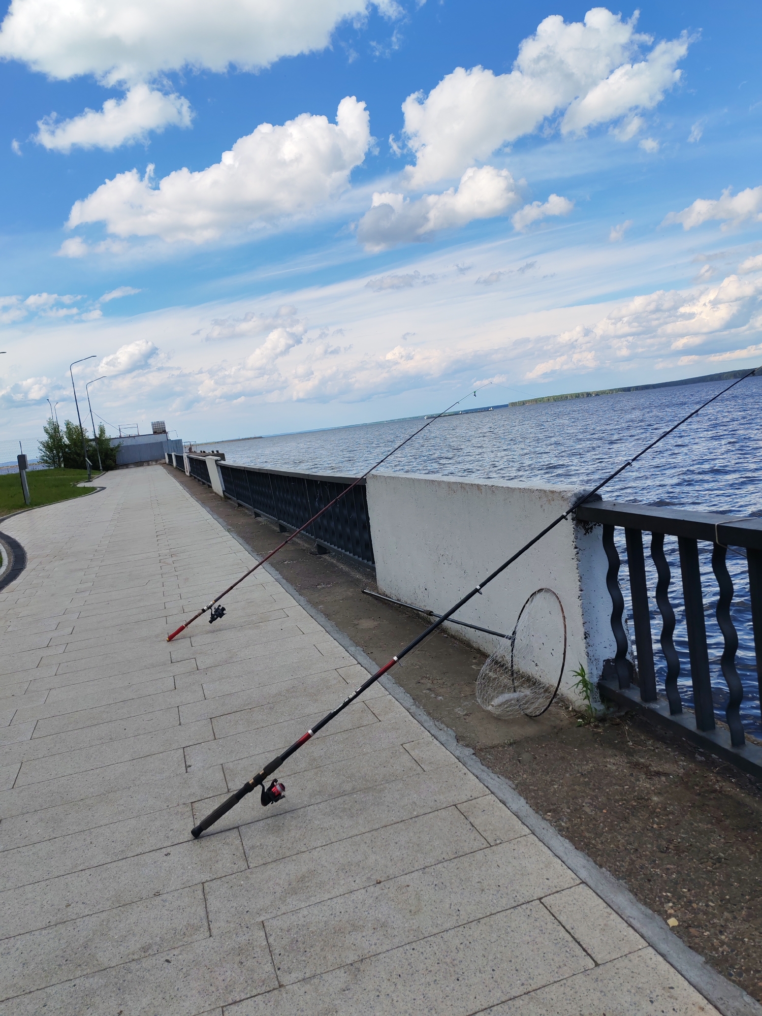 This media file shows a scenic view of a paved promenade along a large body of water, likely a river or lake. Two fishing rods are set up on the promenade, one in the foreground leaning against a concrete barrier and another further back. A fishing net lies on the ground near the foreground rod.
The scene is set under a bright blue sky dotted with fluffy white clouds, suggesting pleasant weather and daytime. The water is calm and blue, reflecting the sky. The promenade itself is paved with large, light-colored tiles and curves gently away from the viewer. A metal railing with an ornate pattern separates the promenade from the water. In the background, the shoreline is visible with some trees and structures.
The presence of fishing rods and a net indicates that fishing is either in progress or has recently occurred. The overall atmosphere is peaceful and recreational, evoking a sense of leisure and connection with nature. The location appears to be a public waterfront area, possibly a park or embankment in Chaikovsky, Russia, given the provided context. There is no visible text in the image.