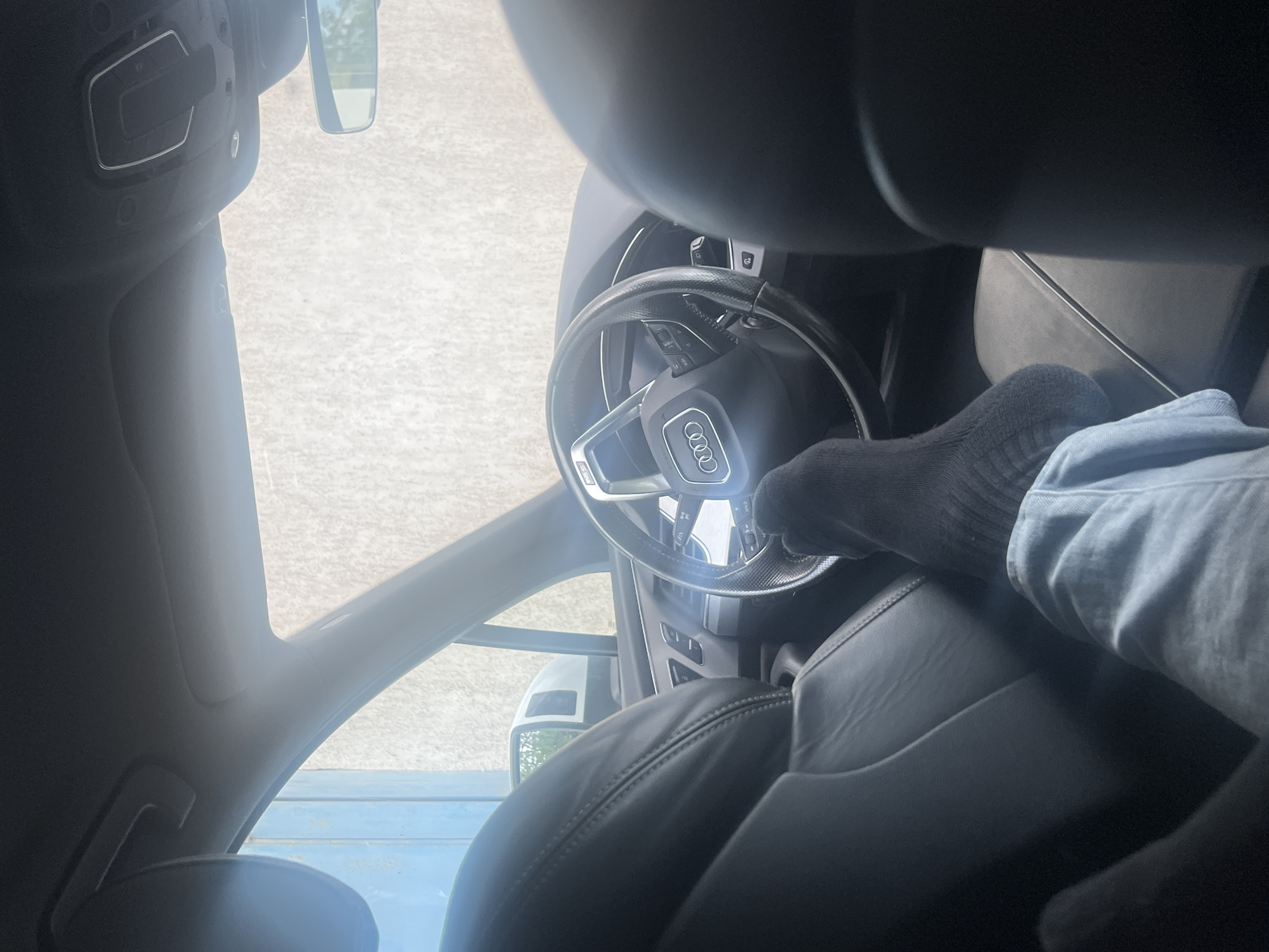 The image depicts the interior of an Audi car, specifically the driver's side. The focus is on the steering wheel, which has the Audi logo prominently displayed. A hand wearing a grey sock and light blue denim is resting on the steering wheel. The car seat is black leather. The car appears to be parked or stationary, as the background is visible through the windshield and side window, showing an outdoor scene, possibly a parking lot. The presence of a rearview mirror confirms the vehicle's interior context. The location cannot be determined to be Varna, Bulgaria from the provided image.