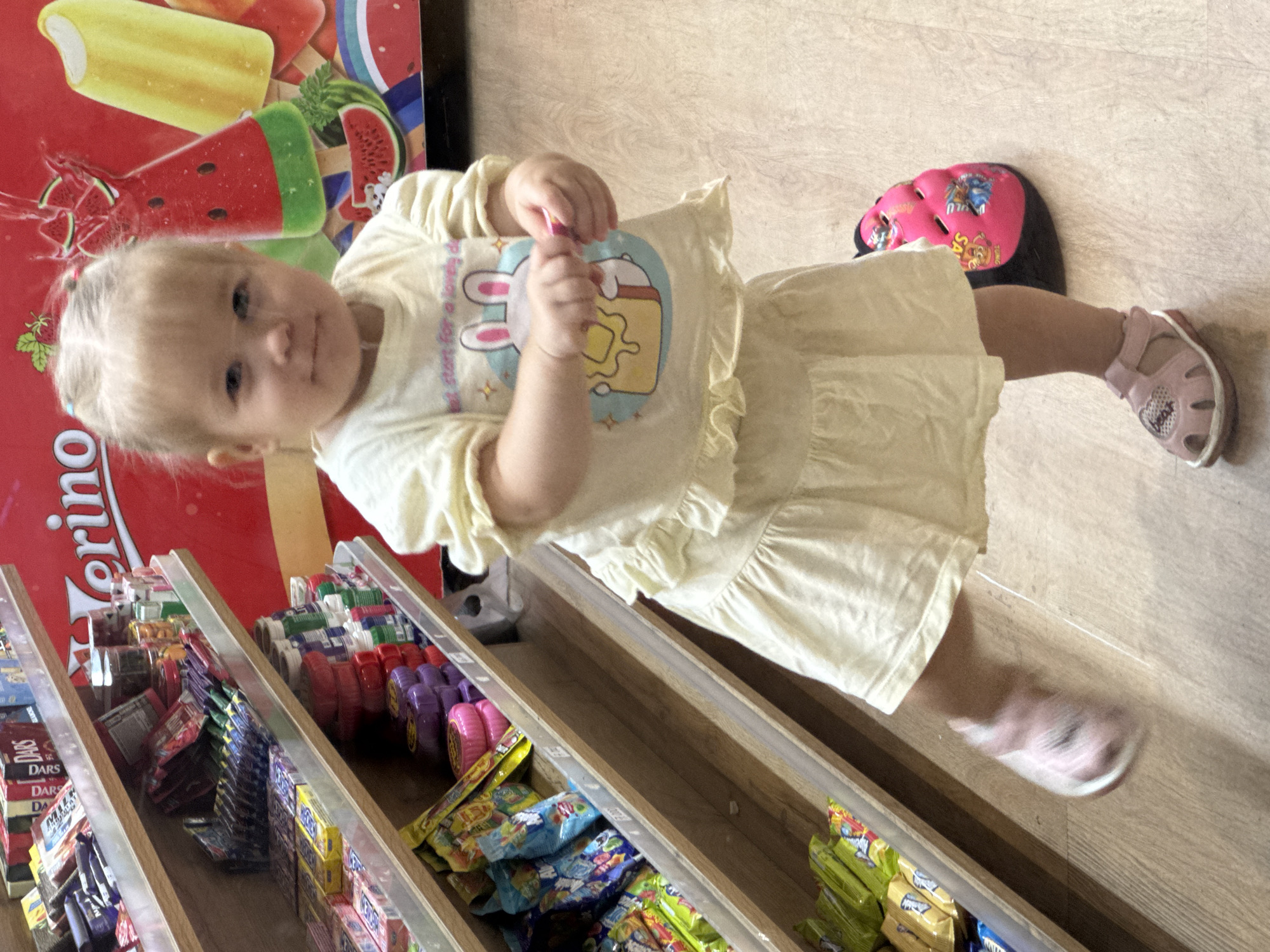 The image shows a young Caucasian girl, approximately 2-3 years old, standing in what appears to be a small convenience store or market. The girl is wearing a light yellow dress with ruffles and a t-shirt with a graphic of a cartoon bunny. She has blonde hair tied in small bunches. In front of her, to her right, is a pink bicycle helmet with graphics of cartoon characters. The girl is looking towards the camera with a neutral expression. She is standing on a wooden floor. Behind her are shelves stocked with various candies and snacks, including packaged candies, small jars, and bubble gum. A large red advertisement with images of ice cream, including watermelon-flavored popsicles, is visible in the background. The store appears to be well-lit, with the bright red advertisement being a prominent feature. The overall impression is that of a casual shopping trip where the child is exploring the store. There is no information in the image to suggest the location is in Nha Trang, Vietnam.