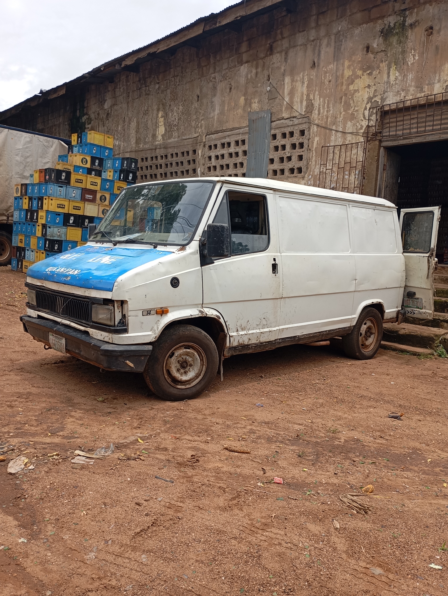 In Shendam, Nigeria, a white panel van with a blue hood is parked on a dirt lot in front of a weathered, aged building. Stacks of blue and yellow crates, some labeled "STAR" and "LEGEND," are piled high to the left of the van, suggesting a distribution or storage area. The van itself appears well-used, with visible wear and tear, including dents and faded paint. Its front is facing the viewer, with the driver's side door on the left and the rear cargo door on the right slightly ajar. The ground is covered in reddish-brown dirt, with scattered debris such as dry leaves, small pieces of plastic, and some organic material. The building behind the van has a corrugated metal roof and a rough, textured concrete or stucco facade with visible signs of age and deterioration, including peeling paint and sections of exposed brickwork. The overall scene depicts an industrial or commercial area, possibly a depot or warehouse.