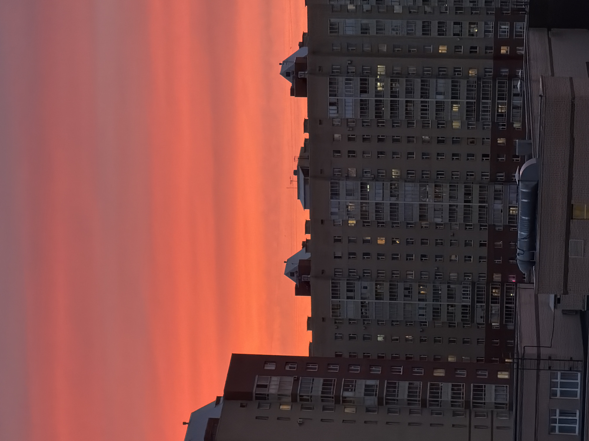 The image depicts a twilight or sunrise scene in an urban environment, most likely in a city. In the foreground are several multi-story apartment buildings with numerous windows. Some of the windows are illuminated, suggesting occupancy. The buildings appear to be modern residential structures. The sky behind the buildings is dominated by shades of orange and pink, indicating either a sunset or sunrise. The lighting suggests that the image was captured during a transition period between day and night. Based on the architecture and urban setting, a possible location for this scene is Saint Petersburg, Russia.