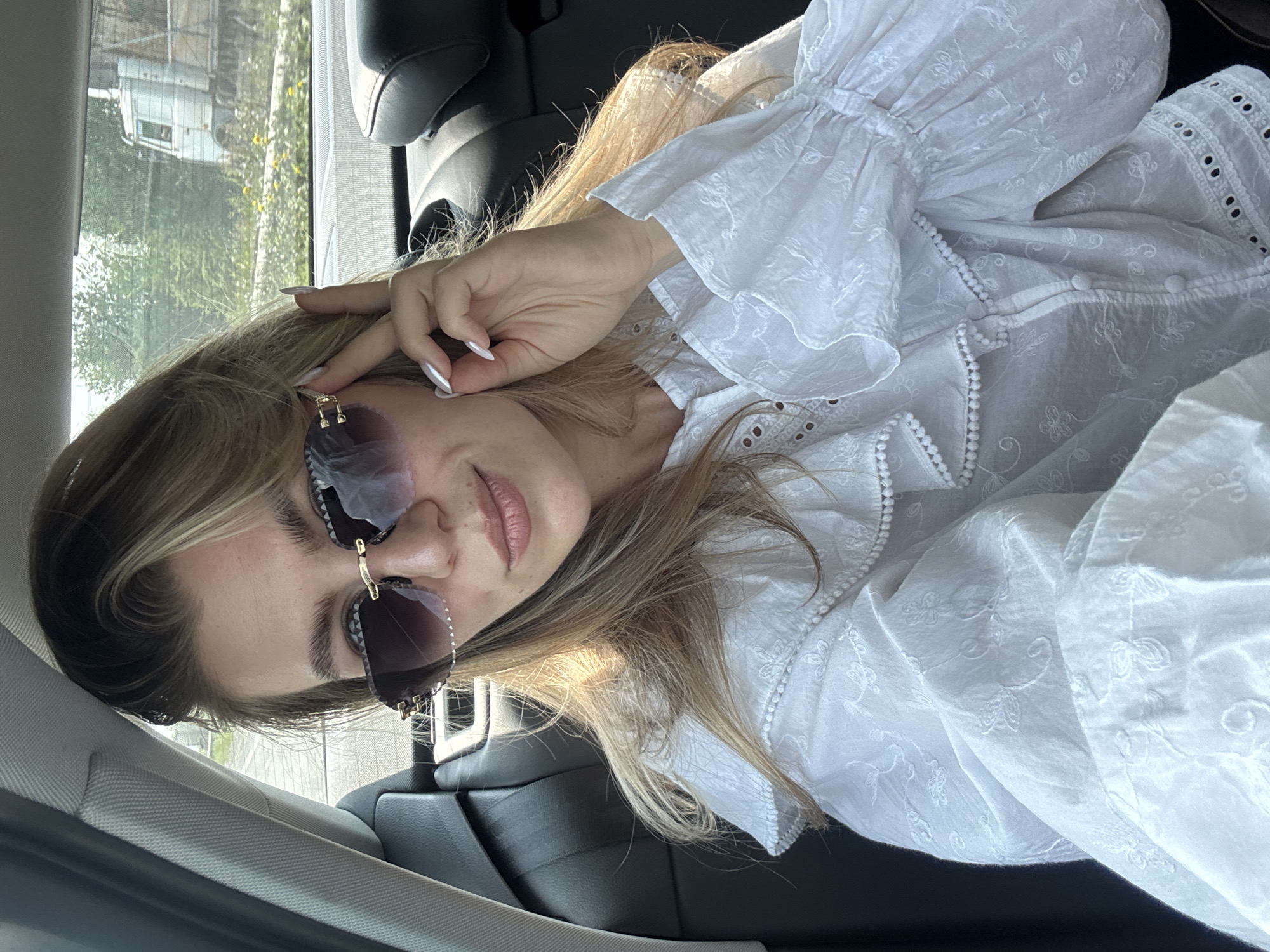 A young woman with blonde hair, wearing sunglasses and a white ruffled blouse, sits in the back seat of a car. She is looking at the camera and adjusting her sunglasses with her right hand. The background shows trees and a truck through the car window.