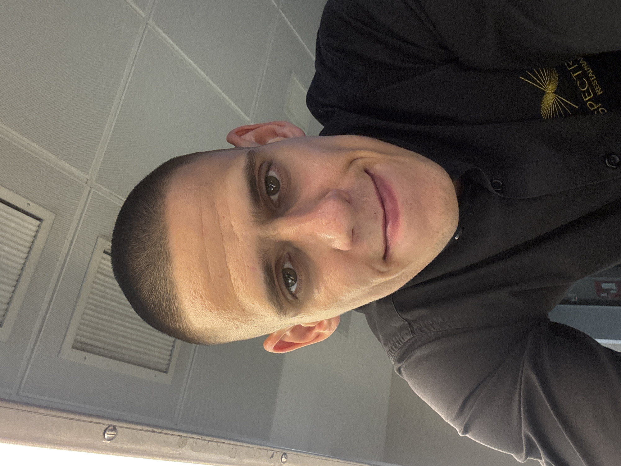 A close-up, low-angle selfie of a young adult male with a buzz cut and olive skin is shown. He is wearing a black collared shirt with a gold embroidered logo that reads "SPECTRA RESTAURANT". The background is an indoor ceiling with fluorescent lighting and a ventilation grate. The man is smiling slightly, and his gaze is directed towards the camera. The photo appears to be taken in a restaurant environment. No specific location in Sofia, Bulgaria is discernible.