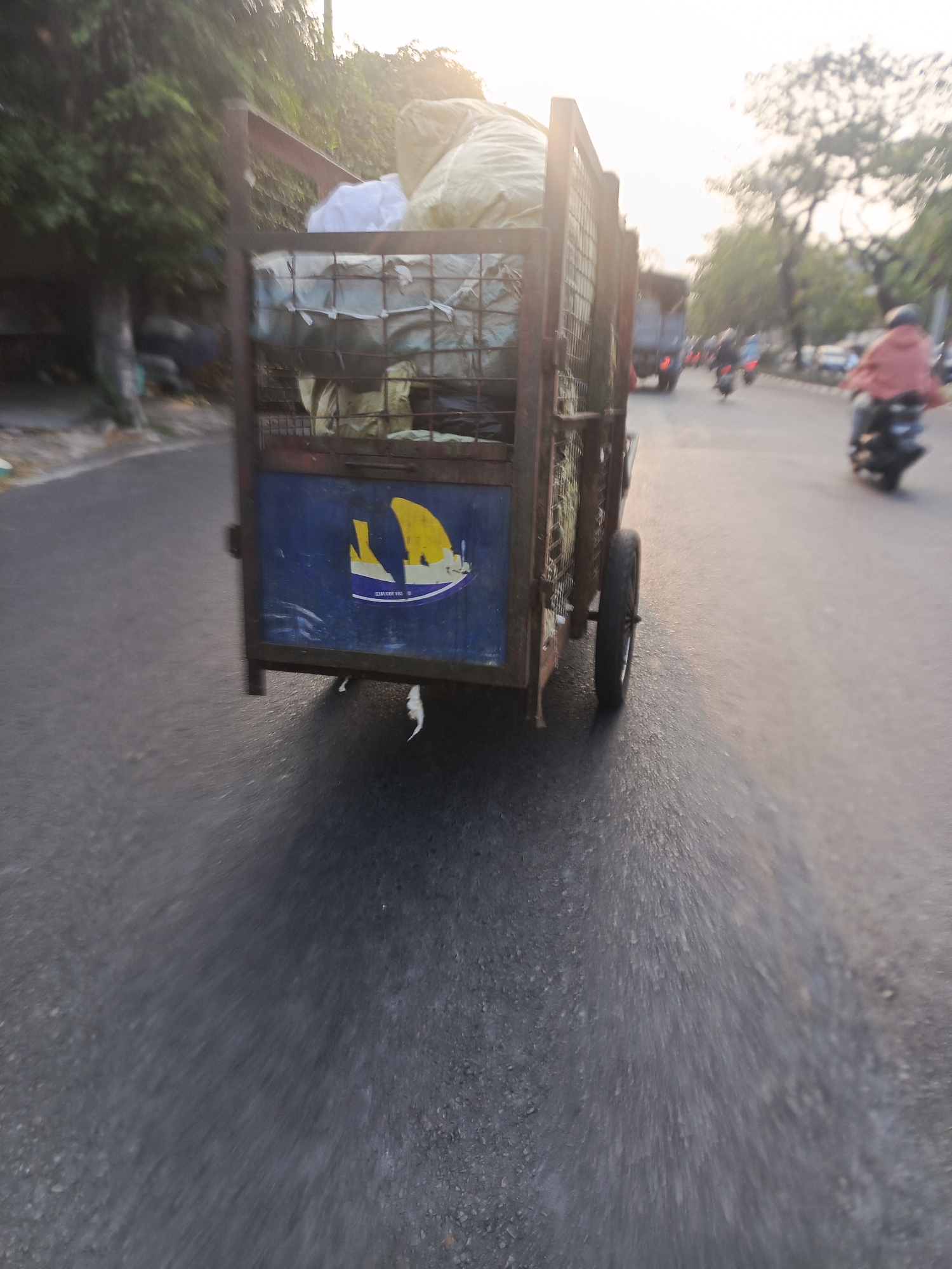 In the city of Surabaya, Indonesia, a street scene unfolds with a focus on a mobile cart laden with goods. The cart, seen from the rear, is a utilitarian structure with a rusted metal frame and a cage-like enclosure, suggesting it is used for transporting items. A blue panel adorns the back of the cart, featuring a stylized yellow and white image of a boat against a blue background, possibly a logo or insignia. The cart is filled with various large bags, some appearing to be fabric or burlap, possibly containing agricultural produce or salvaged materials.
The cart is positioned on a paved road, indicating an urban or semi-urban setting within Surabaya. The road surface is asphalt, and a distinct shadow is cast by the cart, suggesting it is daytime. The motion blur evident in the image implies that the cart is either moving or the photograph was taken from a moving vehicle.
In the background, other elements of a street scene are visible. A person on a motorcycle, wearing a pink garment, is in motion, adding to the sense of activity. Another motorcycle and a truck are also discernible in the distance, suggesting traffic on the road. Trees line the side of the road, with foliage creating a natural backdrop. The sky appears bright, with sunlight filtering through, possibly indicating early morning or late afternoon. The overall impression is a snapshot of daily life and transportation in a bustling Indonesian city.