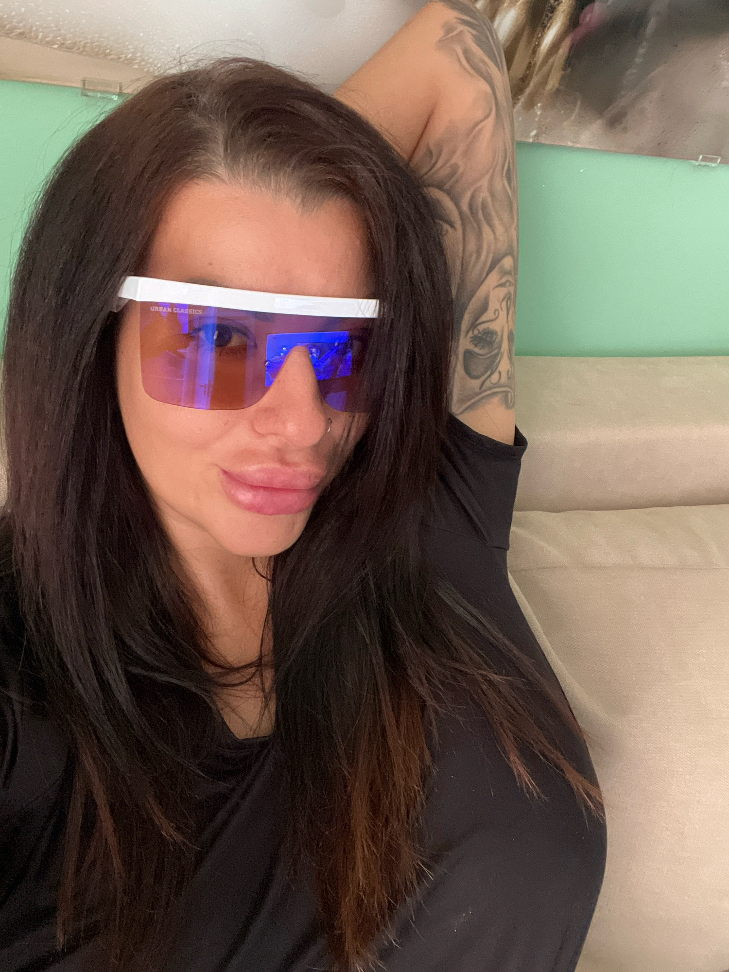 A medium shot captures a woman from the chest up, posed indoors. She has long, dark hair and is wearing large, white-framed sunglasses with pink lenses. A tattoo is visible on her upper right arm. She is wearing a black top. The background consists of a light-colored sofa and a light green wall. The scene appears to be a casual indoor setting. The location is not specified in the image or its metadata.