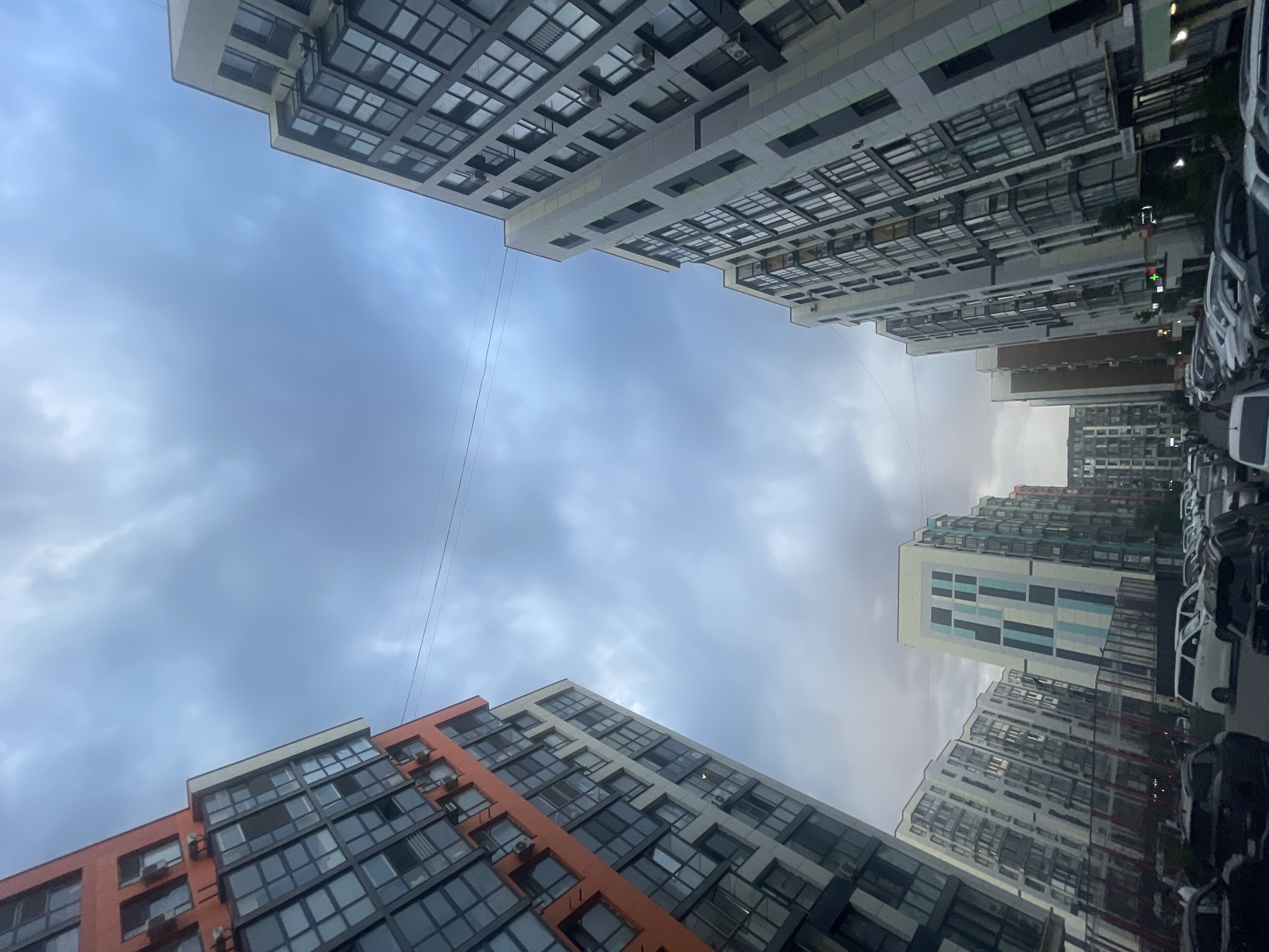 A low-angle, wide shot captures a street scene in Almaty, Kazakhstan, under a cloudy sky. Modern apartment buildings with glass facades and balconies line both sides of the street, which is filled with parked cars. One building on the left is accented with bright orange panels. The buildings exhibit varying architectural styles, with some featuring colorful geometric patterns. The overall impression is of a densely populated, contemporary urban residential area.