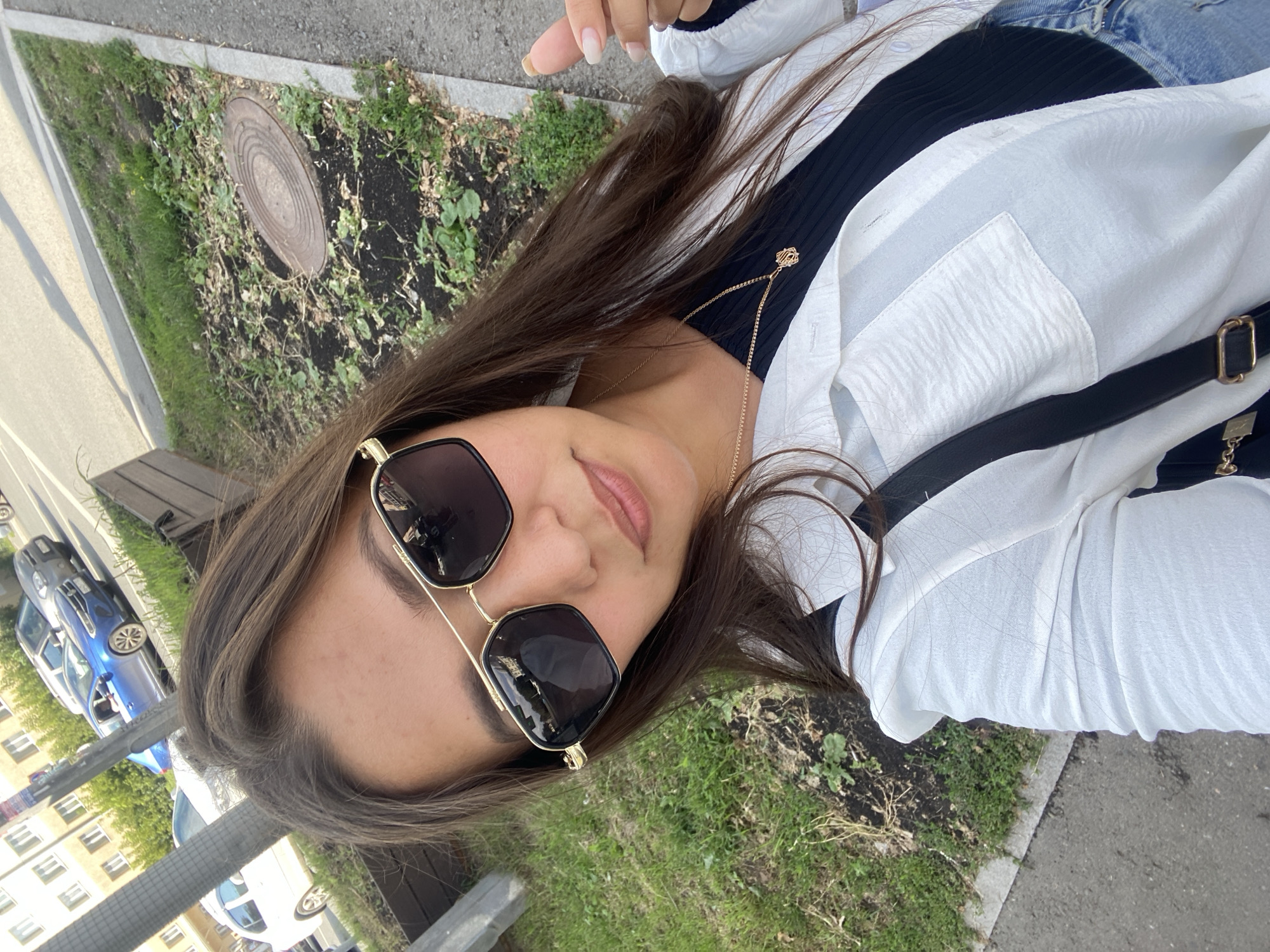 The image shows a young woman taking a selfie outdoors, with a slight tilt to the right. She has long, brown hair and is wearing sunglasses with dark lenses and gold frames. Her outfit consists of a white, loose-fitting shirt over a black ribbed top, and blue jeans. A black strap, possibly from a bag, is visible across her chest. A delicate gold necklace with a geometric pendant is worn over her black top. The background suggests an urban environment, with a patch of grass and dark soil on the left, likely a garden bed with a circular metal grate. Beyond that, a paved area and a street with cars are visible. One car, a blue hatchback, is particularly noticeable. Buildings with windows are visible in the distance. The lighting indicates it is daytime, with sunlight casting shadows. The overall mood appears casual and relaxed. No visible text is present in the image.