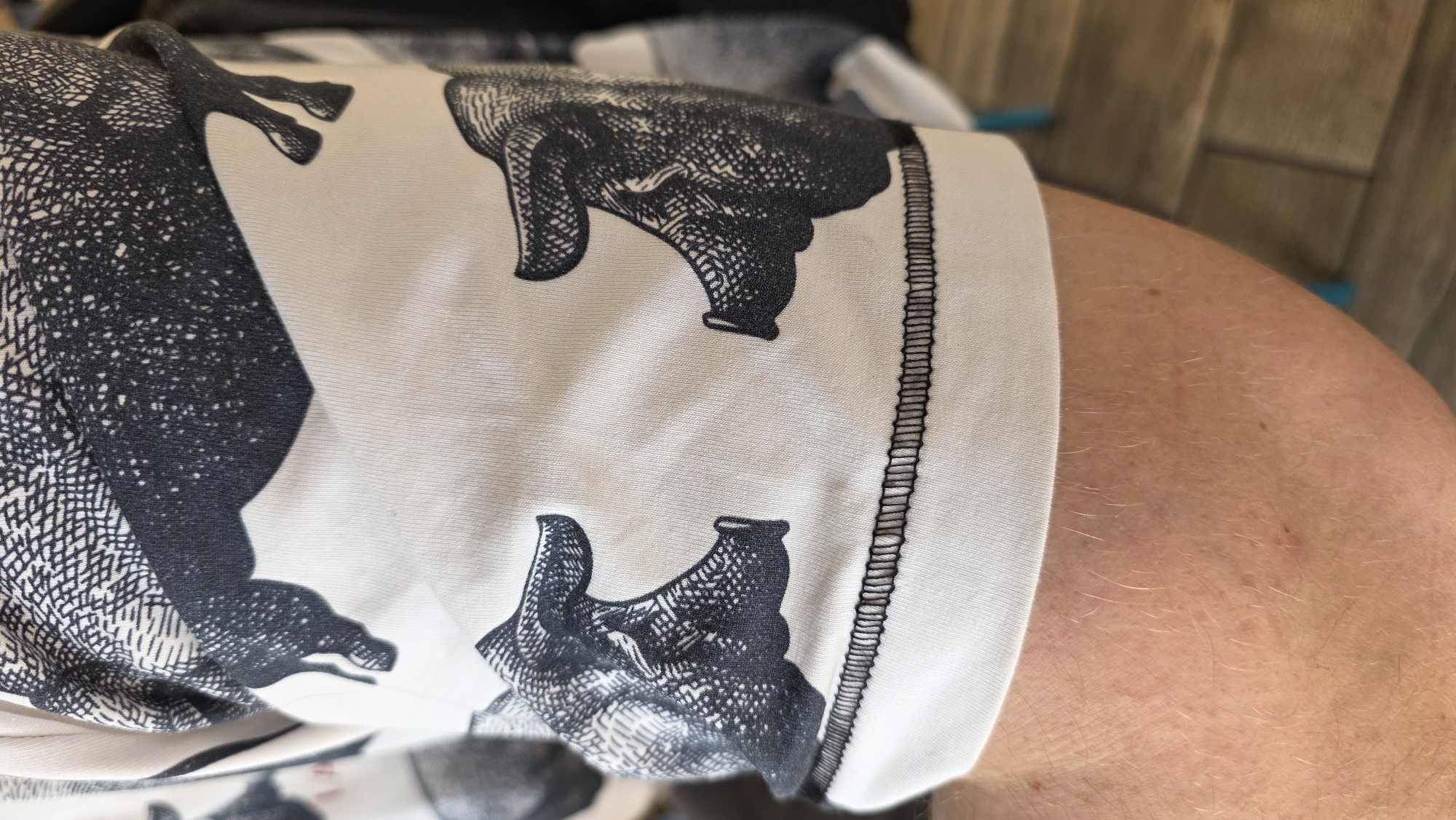 A close-up shot captures a person's lightly tanned arm, adorned with fine, light-colored hair and a few small freckles or moles. The arm is encased in a light-colored garment, likely white or off-white, featuring a striking black and white pattern. The pattern depicts multiple stylized animal figures, resembling pigs or wild boars, rendered in an engraved or woodcut artistic style with detailed cross-hatching and dot patterns providing texture to the dark areas. One animal figure clearly displays a prominent snout-like head. A prominent black stitched seam runs vertically along the right edge of the garment, indicating its construction. The background is softly blurred, showing vertical wooden panels, suggesting an indoor setting with wood-paneled walls or flooring, or an outdoor wooden deck. The context implies the location is Saint Petersburg, Russia. No specific activity or visible text is present.