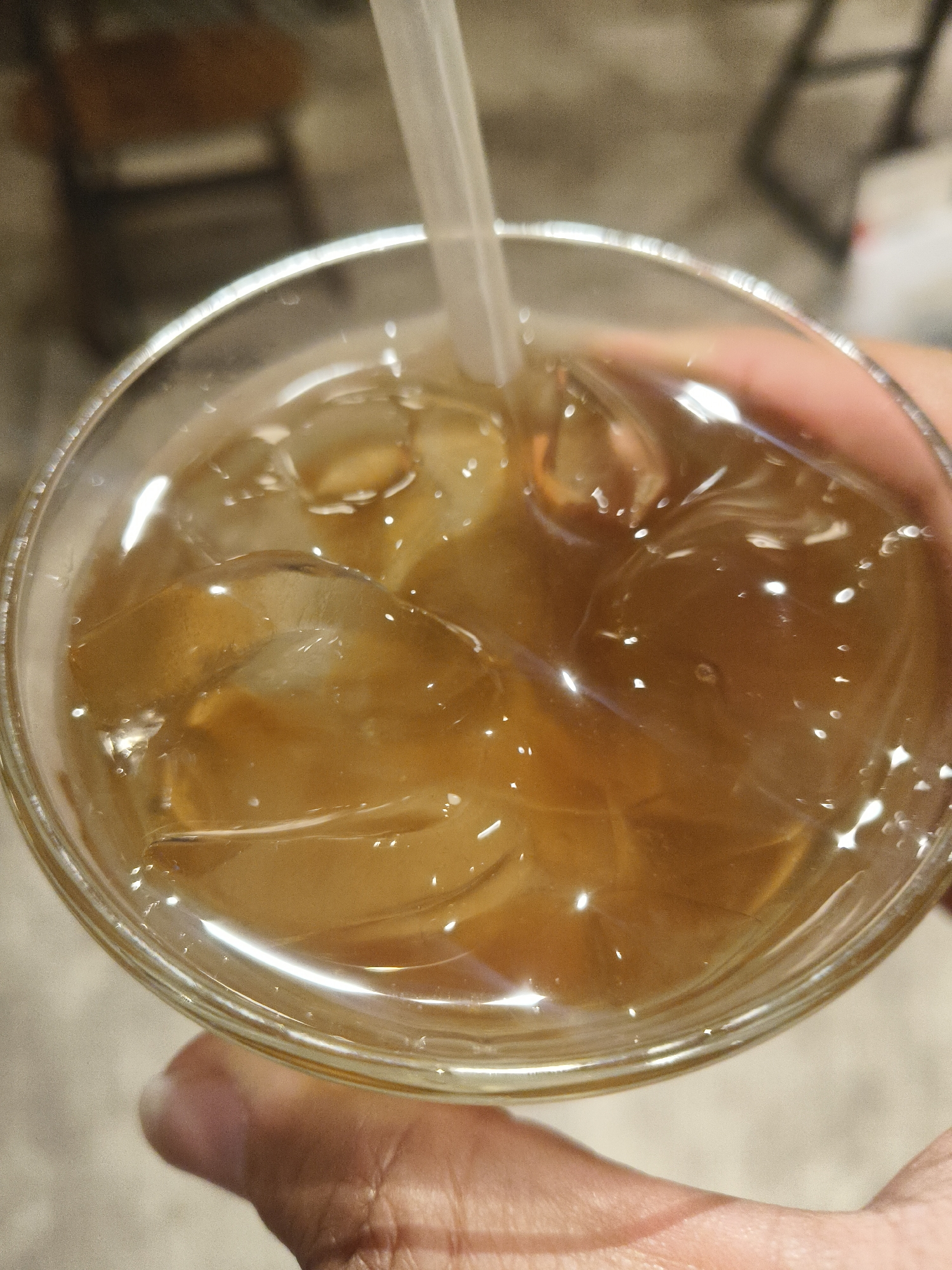 A close-up, overhead shot shows a glass of iced tea being held by a person's hand in Kuta Selatan, Indonesia. The glass is filled with a golden-brown liquid and numerous ice cubes. A white plastic straw is visible submerged in the drink. The background is blurred, but suggests an indoor setting with hints of furniture. The lighting is bright, causing reflections on the ice and the surface of the liquid.