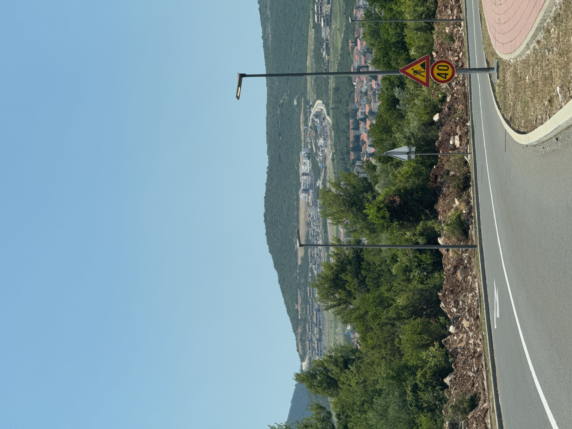 The image shows a road curving to the right in Đuraševići, Montenegro. On the side of the road, there are two traffic signs. The upper sign is a yellow triangle with a red border, depicting a person digging. Below it, a circular red sign with a white border displays the number "40" in black, indicating a speed limit. Further down the road, a street lamp is visible. In the background, lush green trees dominate the landscape, with a scattering of buildings on a hillside under a clear blue sky. The road itself is asphalt with a white line marking the lane. The surrounding area features a mixture of dry grass, dirt, and rocks, suggesting a rural or semi-developed environment.