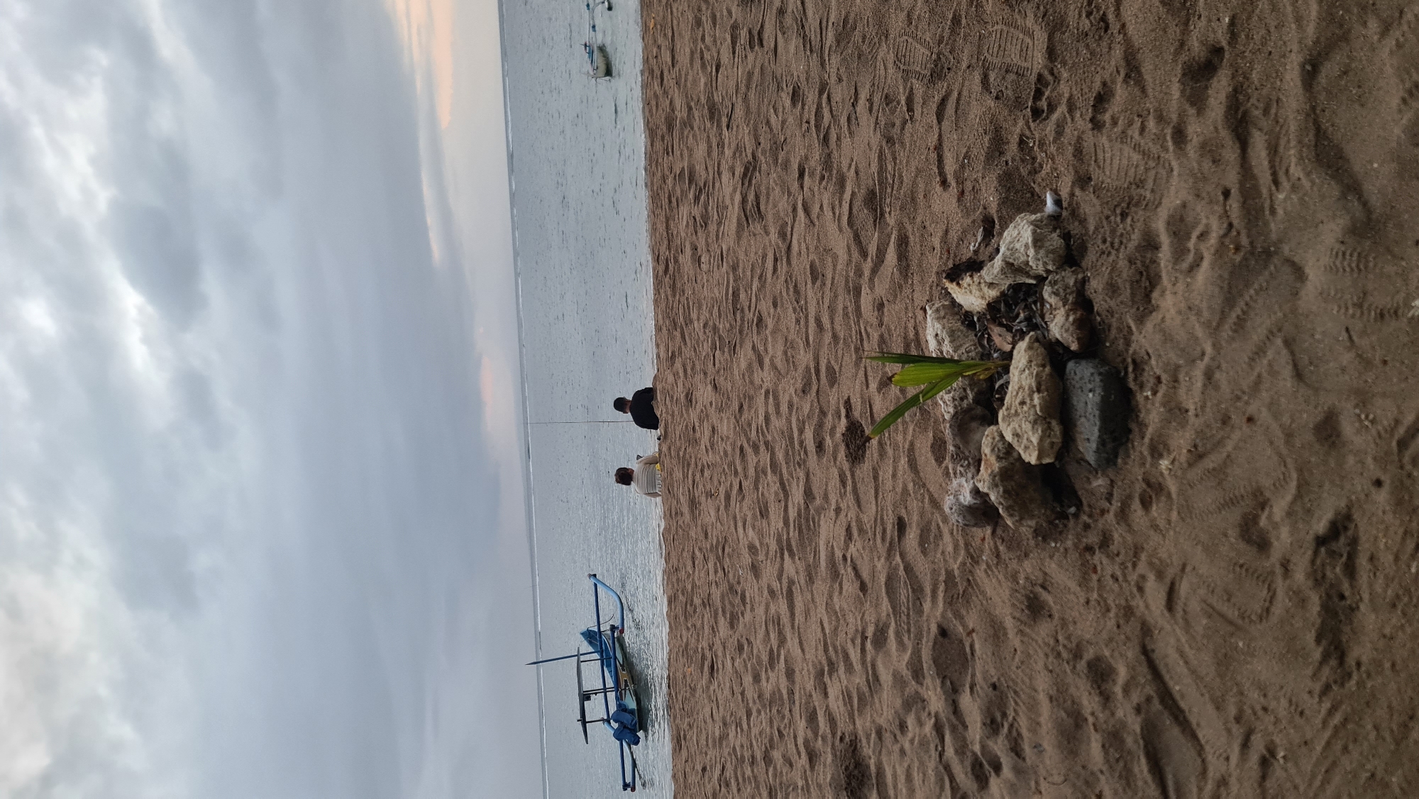 The image depicts a beach scene at dusk on the coast of Denpasar, Indonesia. The foreground is dominated by sand, with a small arrangement of rocks and a single plant sprout in the center. In the middle ground, two individuals are seated on the sand facing the water. One person is wearing a light-colored shirt, and the other is wearing a dark shirt. They appear to be engaged in fishing, as a fishing rod is visible behind them. To the left, a traditional Balinese fishing boat with outriggers is moored in the calm sea. The sea extends to the horizon, where it meets a cloudy sky with hints of soft, warm light suggesting the setting sun. Another small boat is visible in the distance to the right. The overall atmosphere is tranquil and serene, indicative of a peaceful evening by the ocean.