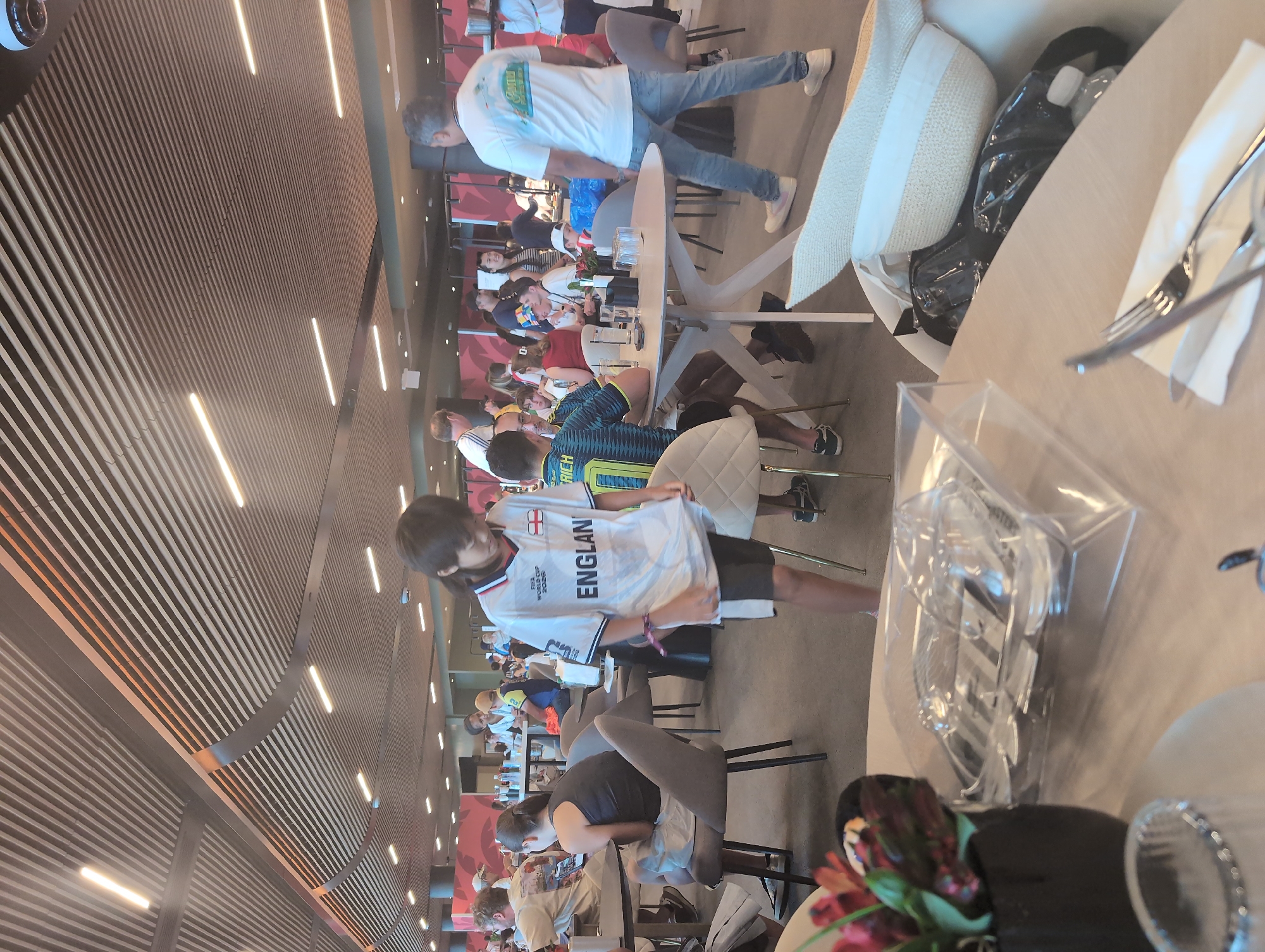 In a brightly lit indoor seating area, likely a stadium concourse or restaurant, at the Hard Rock Stadium in Miami Gardens, Florida, people are casually seated at tables. A young person wearing an England soccer jersey stands in the foreground, looking down. To the left, a person in a dark tank top sits at a table, looking at a phone. To the right, a man in a white t-shirt and jeans walks away from the camera. In the background, various groups of people are seated at tables, some interacting with each other, others engaged with their phones. The environment features modern architecture with slatted ceilings and strategically placed lighting. On the tables, items like glasses, cutlery, and food trays are visible.