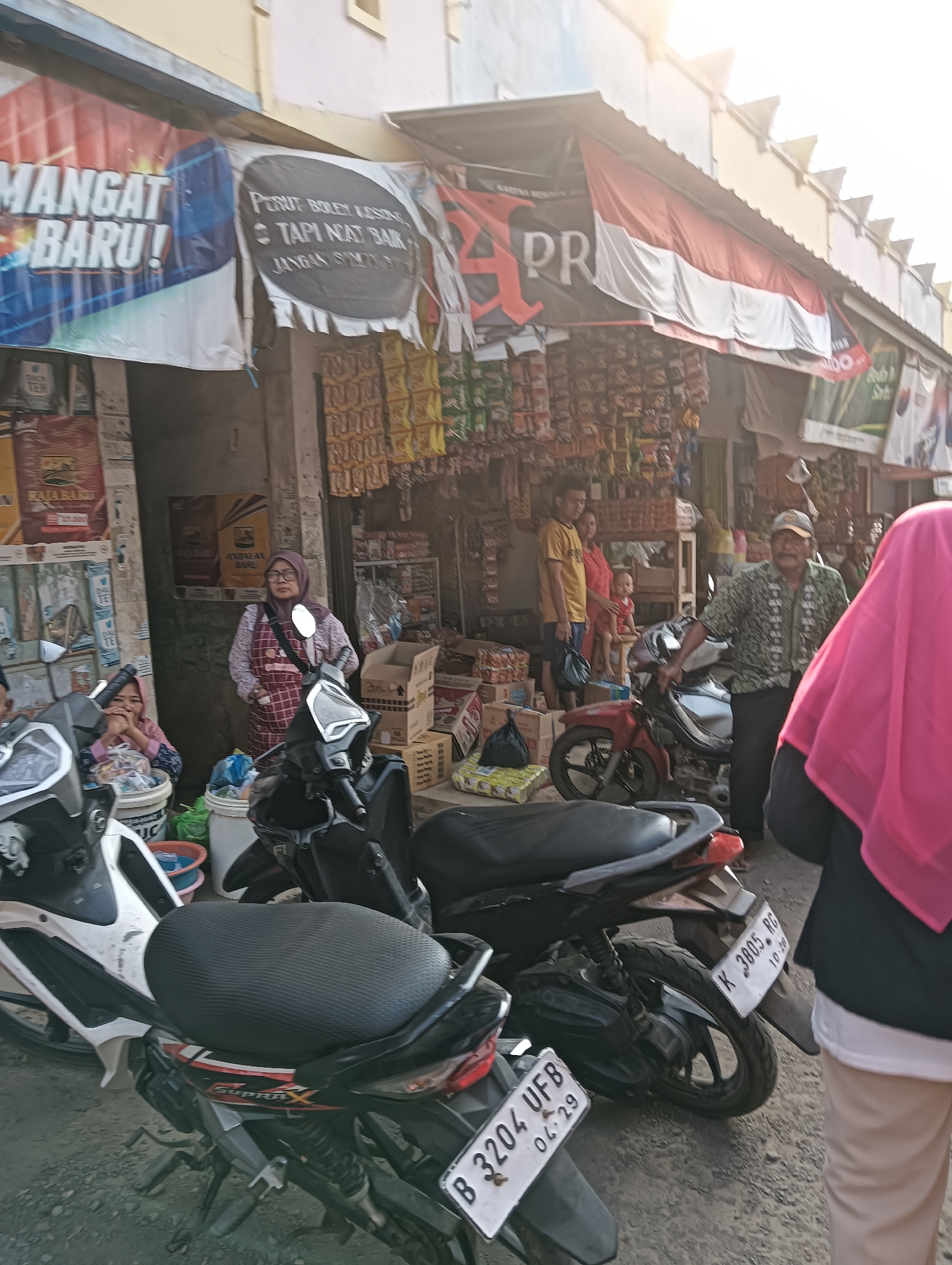 The image shows a street market scene in Cluwak, Indonesia, with several people and motorbikes. In the foreground, there are two motorbikes. The one on the right is black and has a license plate that reads "K 3885 RC 10.20". The one on the left is white and black, and its license plate is partially visible, reading "B 3204 UFB 04.29".
In the middle ground, a man wearing a patterned shirt and a cap is standing next to a red motorbike. To his left, a woman in a red outfit is holding a baby. Further left, a woman wearing a headscarf and a checkered apron is standing near some buckets and plastic bags. Behind them, a street market stall is visible, with various goods displayed on shelves. A banner with the text "MANGAT BARU!" is hanging on the left side of the stall.
In the background, more market stalls and buildings are visible, along with a large Indonesian flag hanging across the street. The overall environment appears to be an outdoor market on a bright, sunny day. The ground is covered in dirt and gravel.