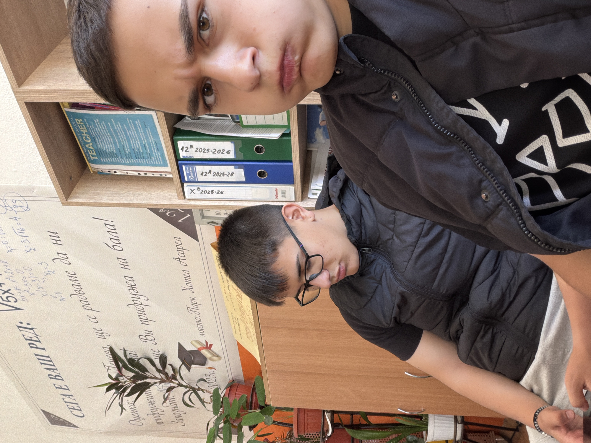 Two male individuals are present indoors. The first individual is seen from the head up in the top portion of the frame with an annoyed expression. The second individual appears from the chest up and is wearing glasses and looking down. Both individuals are wearing black outerwear. A poster on the wall reads "SEGA E VASH RED" and contains mathematical equations. It includes the phrase "ще се радваме да ни придружа на бала!" which translates to "We will be happy to accompany you to the ball!" It also references a location, "място: Парк Хотел Асарел," indicating the Park Hotel Asarel as the location in Panagyurishte, Bulgaria. There is a shelf containing books and file folders to the left of the first individual. There are plants near the poster on the bottom left of the frame.