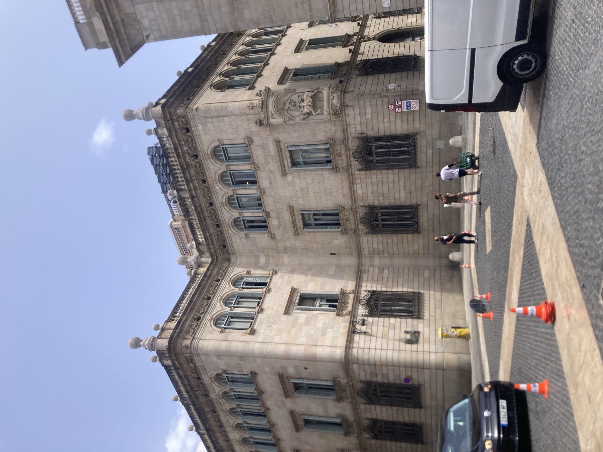 The image shows a street scene in Barcelona, Spain, in front of a large, ornate stone building. The building has multiple stories with arched windows, decorative cornices, and statues. There are several traffic cones on the cobblestone street, suggesting ongoing work or a restricted area. A black car is parked on the left, and a white van is parked on the right. Three people are visible on the street; one pushing a cart and two walking. The sky is clear and blue with a few faint clouds.