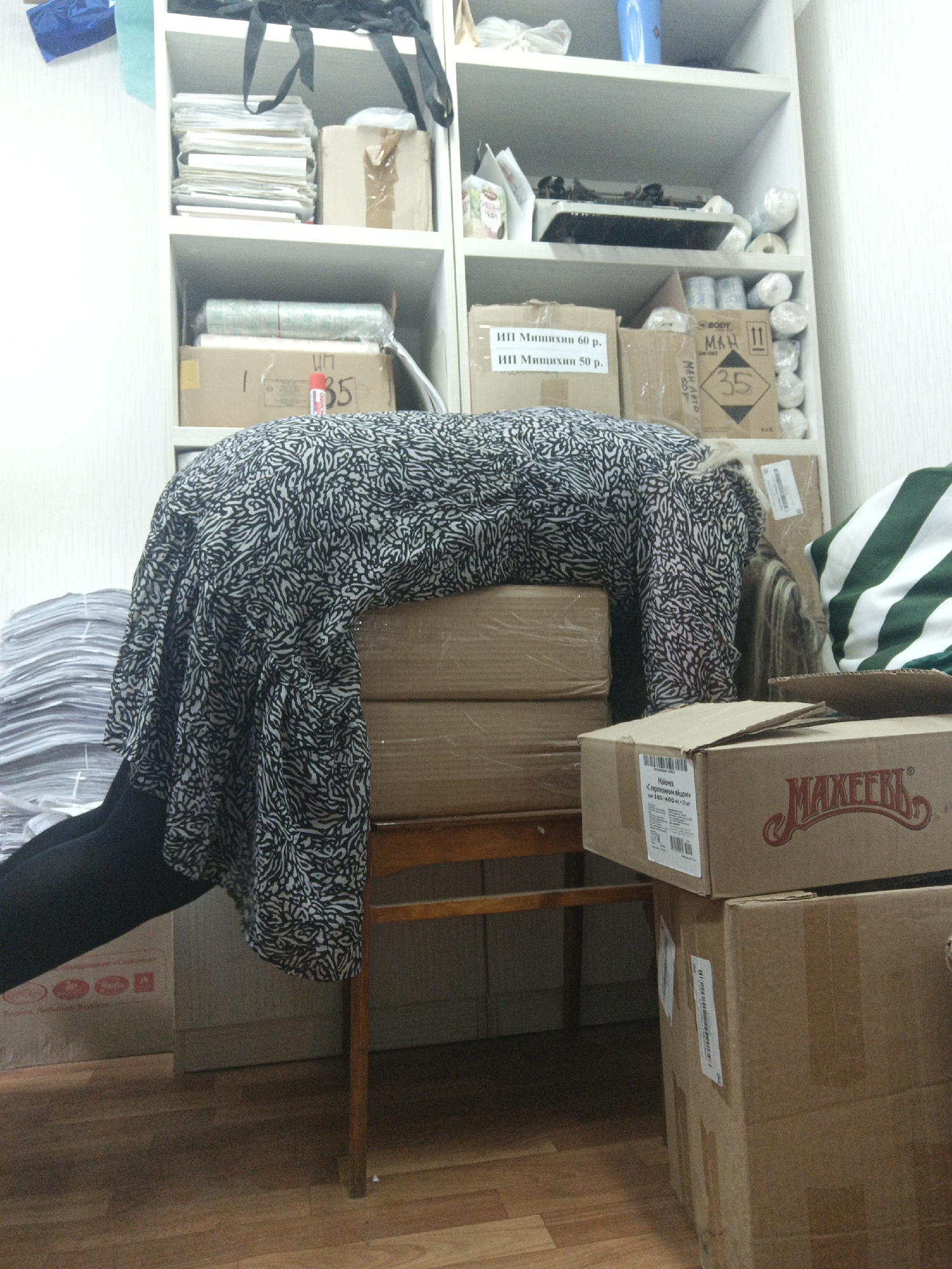 The image depicts an indoor scene, likely a cluttered office or storage room in Noviy, Russia. A person is prominently featured, lying face down across a stack of cardboard boxes, with their upper body covered by a black and white abstract-patterned dress or tunic. Their legs, clad in black tights or pants, extend to the left, and light-colored hair is visible at the back of their head. The boxes they are resting on are supported by a wooden chair.
The background consists of white shelving units filled with various items, including stacks of papers, smaller cardboard boxes, rolls of materials, a blue cup, and a small electronic device. More cardboard boxes are stacked on the floor. To the left, a large pile of documents or papers rests on the floor. A striped green and white cloth or bag is visible on the right side of the frame. The floor is a light-colored wooden laminate.
Visible text on the boxes includes "ИП Мишихин 60 р." and "ИП Мишихин 50 р." (indicating an individual entrepreneur and prices in rubles), "BODY," "МАН 11," and the number "35" which appears on multiple cartons. A brown box in the foreground on the right features the brand "МАХЕЕВЪ" and text reading "Майонез С перепелиным яйцом" (Mayonnaise with quail egg). The person's posture suggests a moment of rest or exhaustion in what appears to be a busy or disorganized workspace.