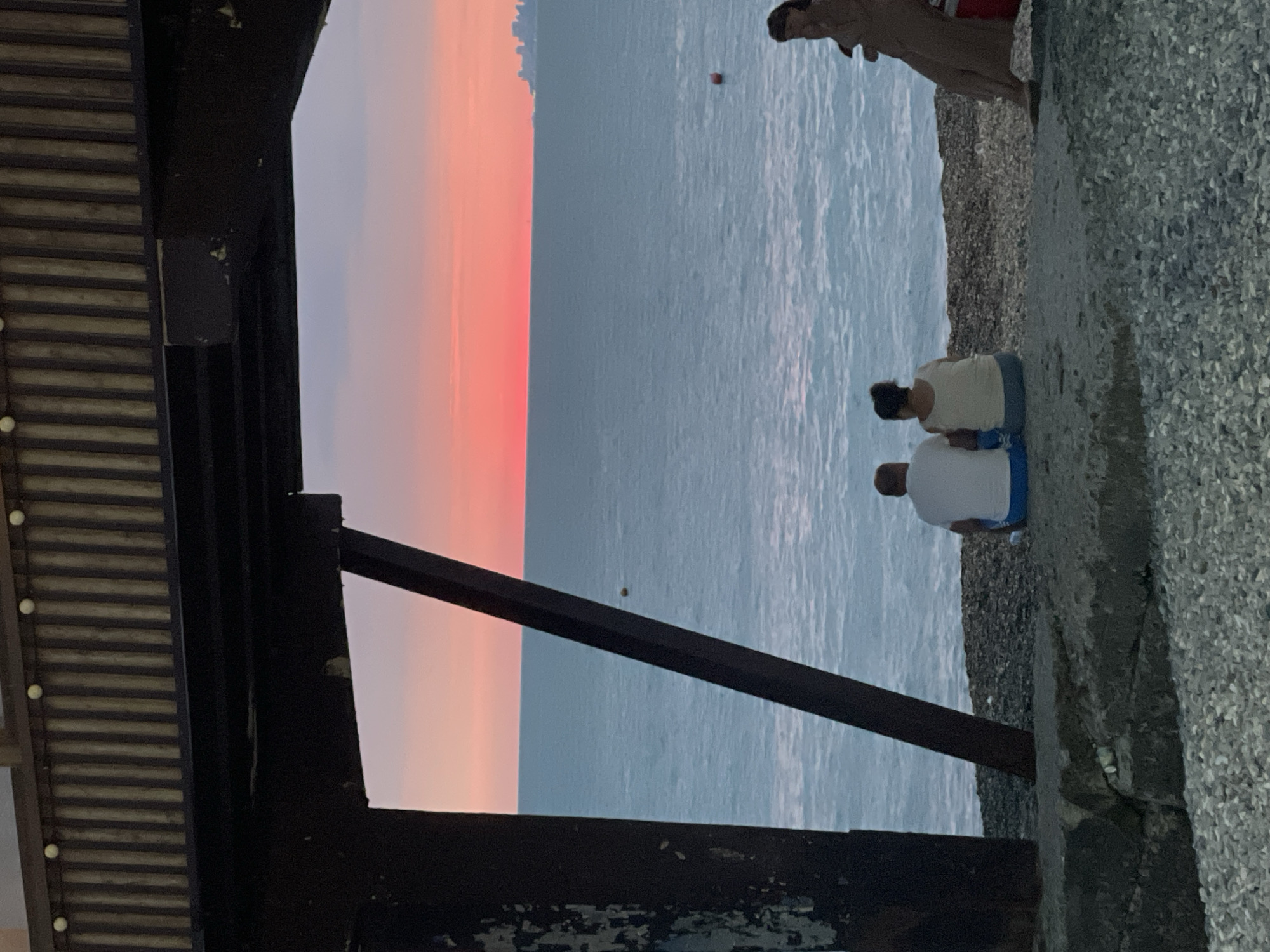 In the city of Unknown, Unknown, the image captures a serene sunset over a tranquil body of water. The sky is painted with vibrant hues of pink and orange, transitioning to a soft lavender as it meets the horizon. The water, a calm expanse of blue-gray, gently laps against a pebbled shore.
In the foreground, a dark, weathered structure, possibly part of a pier or bridge, casts a shadow, framing the scene. Beneath this structure, two individuals are seated on the shore, facing the water and the stunning sunset. The person on the left is wearing a white t-shirt and blue shorts, while the person on the right is in a light-colored tank top and dark pants. Their posture suggests a shared moment of quiet contemplation.
Further to the right, another person is partially visible, reclining on the shore, also facing the water. This individual is wearing a light brown garment and appears relaxed. The presence of buoys in the water indicates it is a frequented area, perhaps a beach or a harbor. The overall atmosphere is one of peacefulness and natural beauty, with the human figures adding a touch of human presence to the expansive landscape.