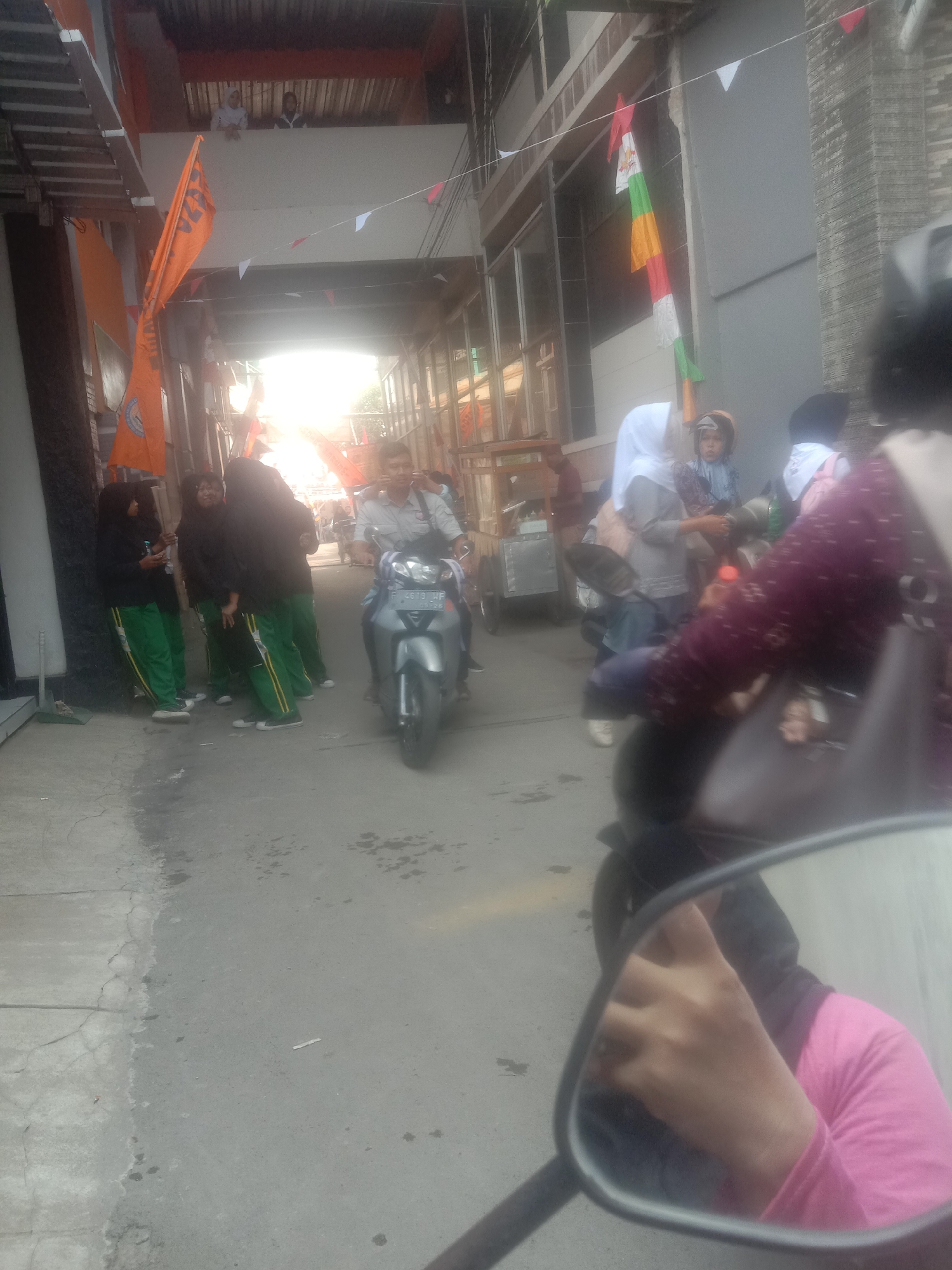 The image depicts a street scene in Karangtengah, Indonesia, likely during daylight hours with bright sunlight illuminating the area. The foreground shows a motorcycle mirror reflecting a person's hand wearing a pink long-sleeved shirt. The road surface is paved and appears somewhat weathered, with some darker patches and cracks.
In the mid-ground, a man is riding a silver motorcycle. He is wearing a light-colored shirt and dark pants. Behind him, a group of four people, appearing to be young women or girls, are standing on the side of the road. They are all wearing black headscarves and green pants with yellow stripes. They seem to be engaged in conversation. Further down the street, other individuals and vehicles are visible, suggesting a moderately busy urban environment.
The background features buildings with corrugated metal roofing and balconies. Orange and white triangular flags are strung across the street, along with some vertical banners. A food cart is also visible to the left. The overall impression is of a lively street in a residential or commercial area.