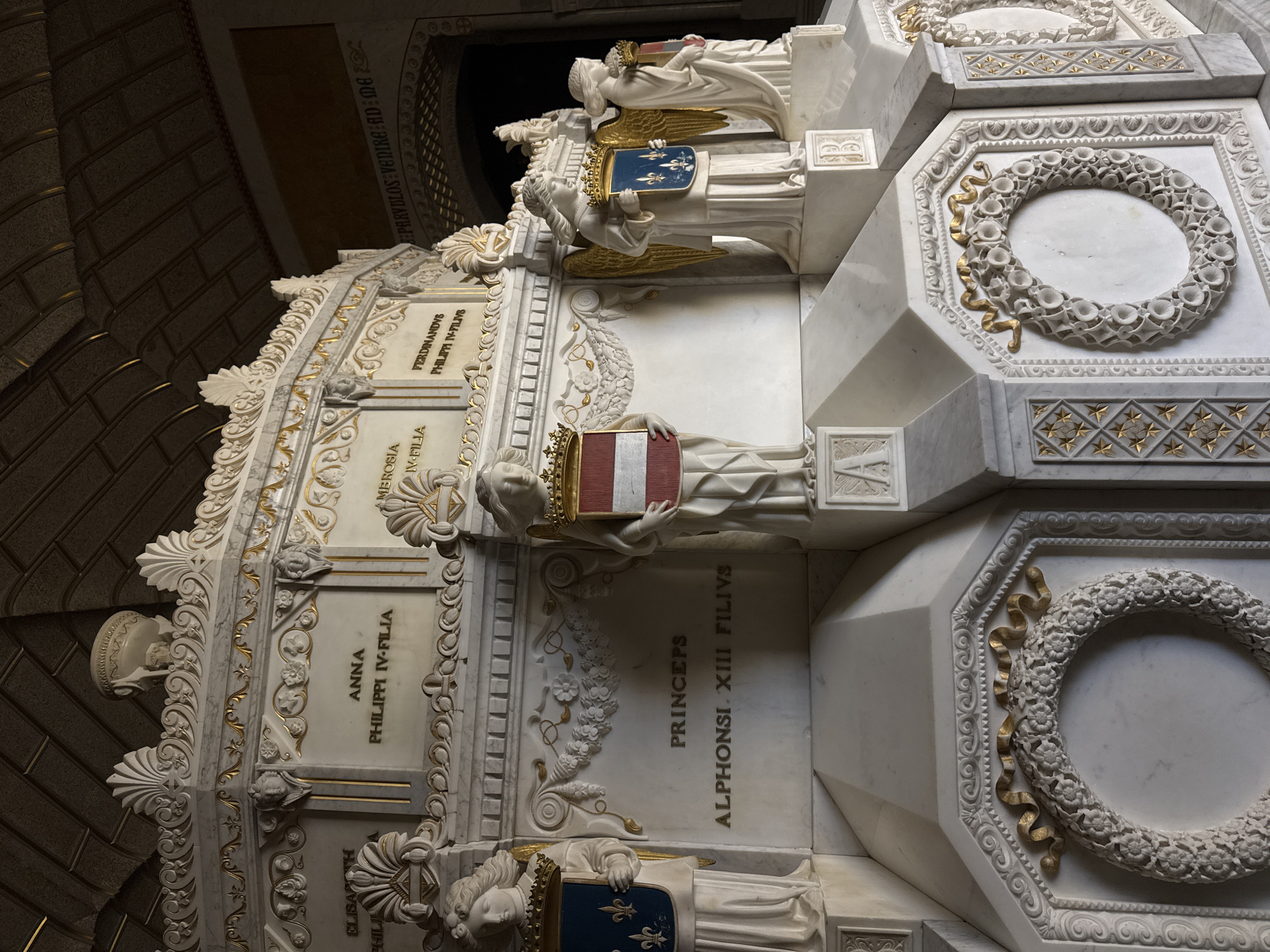 This is a detail of a marble mausoleum in the Basilica of the Monastery of San Lorenzo de El Escorial, Spain. The mausoleum is ornate, featuring white marble with gold accents. It is adorned with carvings of shells, floral wreaths, and cherubic figures. Several marble statues of winged figures, each holding a shield with heraldic symbols, flank the central inscription. One shield displays a blue background with three golden fleur-de-lis, another shows red and white stripes, and a third has a red and gold shield with blue elements. Above the statues, marble plaques bear names and inscriptions. Prominently displayed is the inscription "PRINCEPS ALPHONSI. XIII FILIVS." The ceiling above is arched and appears to be coffered with gold elements. The overall impression is one of grandeur and historical significance.