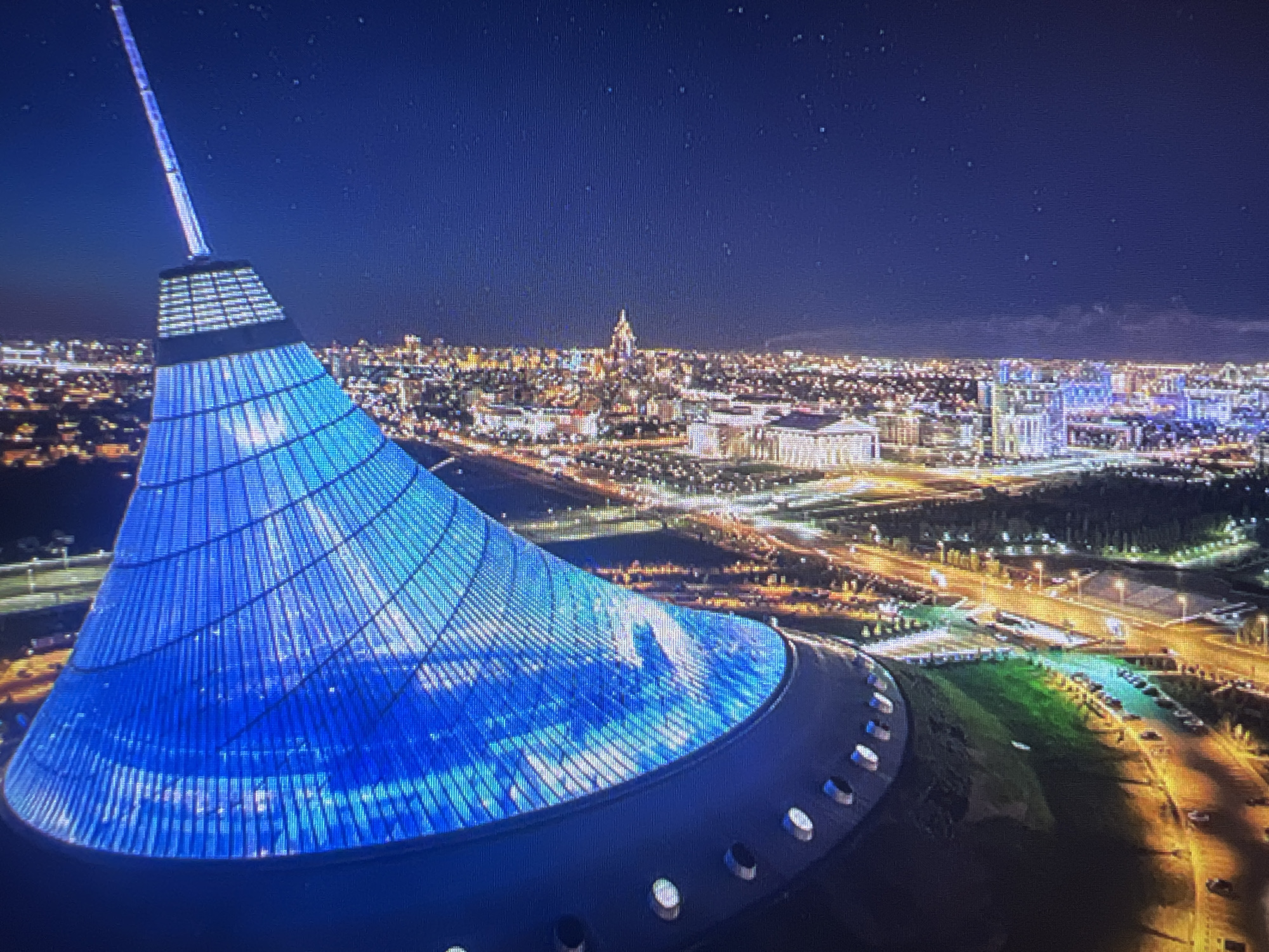 The image captures a nighttime aerial view of the city of Astana, Kazakhstan, with the Bayterek Tower, a prominent landmark, in the foreground. The tower, a cone-shaped structure with a pointed spire, is illuminated with a vibrant blue light, reflecting the night sky. The city unfolds behind it, a tapestry of illuminated buildings, streets, and lights, suggesting a bustling urban environment. The sky above is dark blue, dotted with faint stars. The scene depicts an urban landscape at night, showcasing architectural elements and city lights.