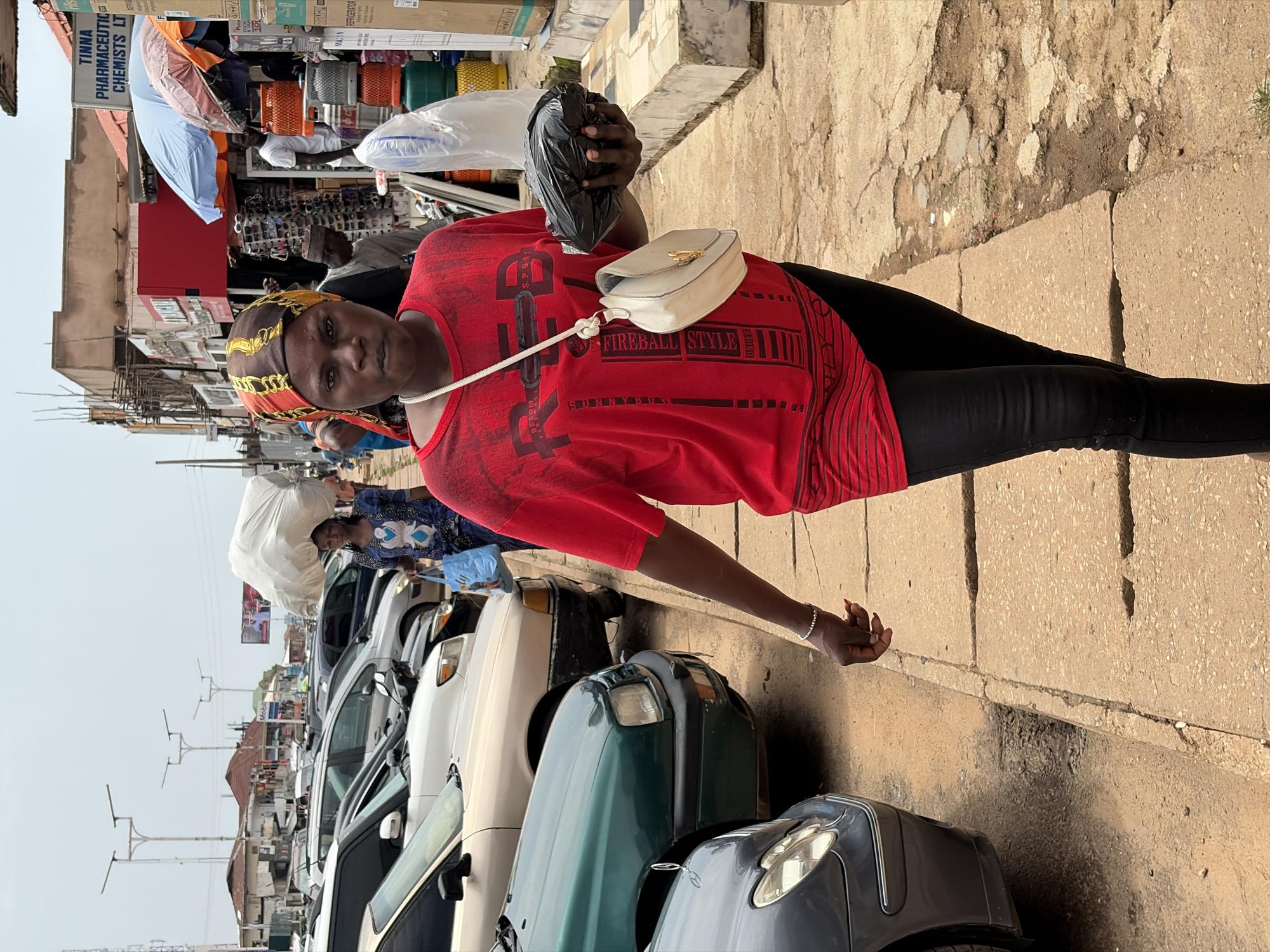 A woman wearing a red t-shirt and black leggings stands on a paved sidewalk. She has a yellow and red patterned headscarf. A white purse with a gold clasp hangs from her shoulder. In her right hand, she holds a black plastic bag. Behind her, a row of cars is parked along the street. In the background, shops line the street, with awnings and various goods displayed. One shop sign reads "TINNA PHARMACEUTIC CHEMISTS LTD." The scene appears to be in a market or busy street in Jos, Nigeria, possibly in the afternoon, judging by the lighting. The woman has a neutral expression.