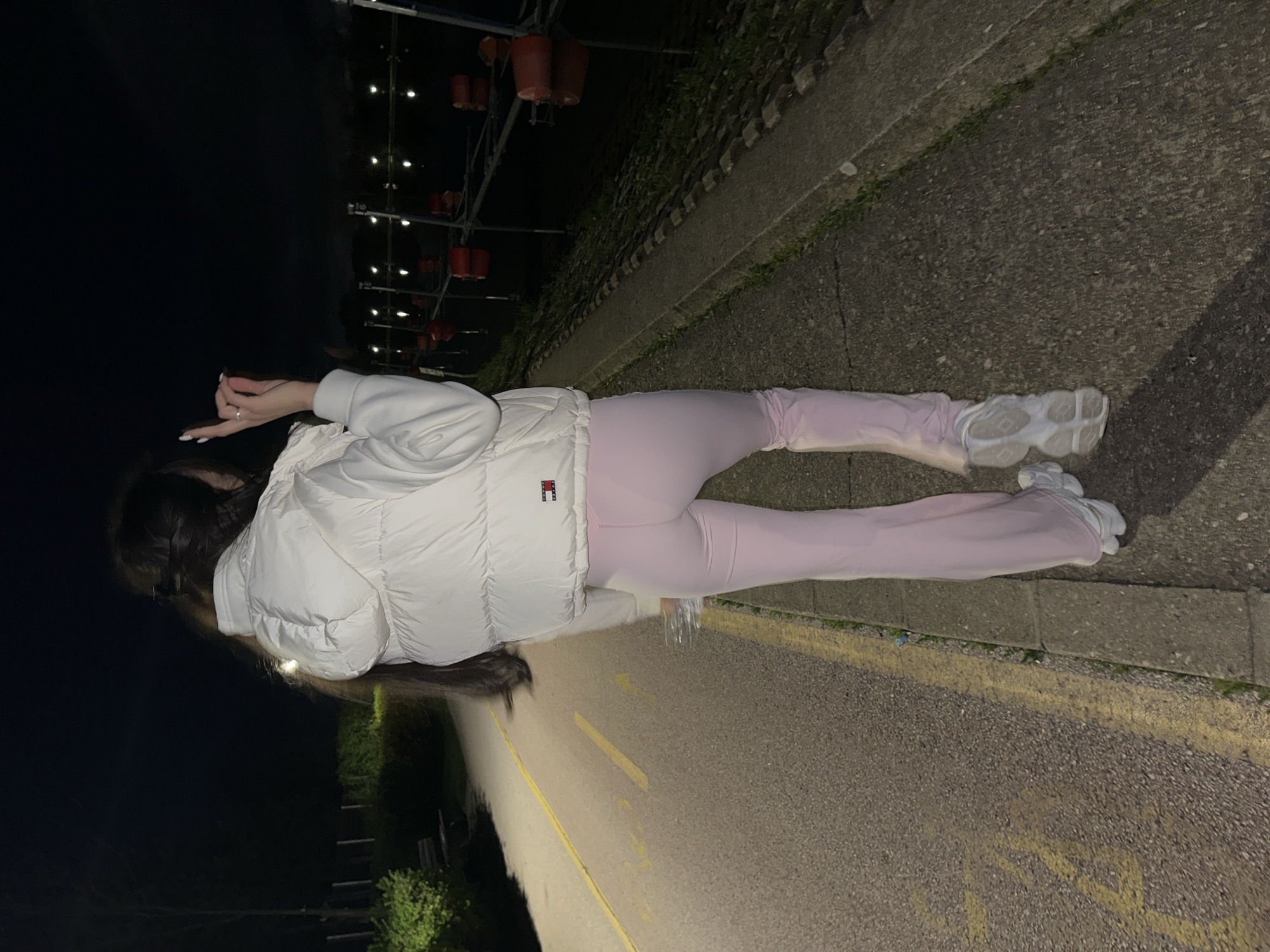 A woman is walking on a paved road in Plovdiv, Bulgaria, at night. She is wearing a white puffer jacket, pink leggings, and white sneakers. The road is next to a concrete wall with vegetation growing on it. There are decorative lanterns hanging overhead.