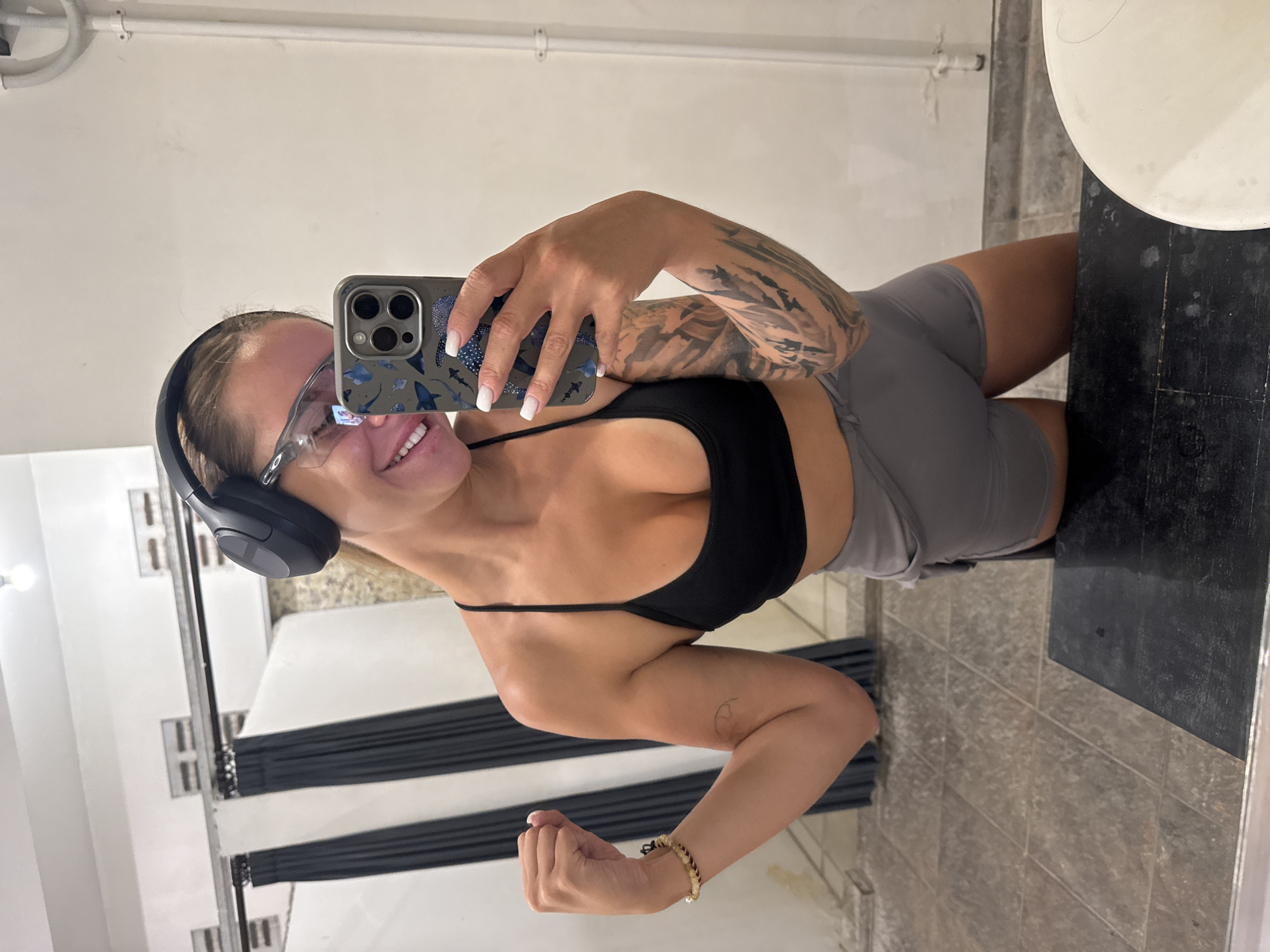 A woman is taking a selfie indoors. She is wearing a black sports bra, gray shorts, headphones, and glasses. Her phone has a blue patterned case. She has tattoos on her left arm and is wearing a bracelet on her right wrist. The location appears to be a bathroom with a sink and striped black and white walls. The environment seems modern. The scene is consistent with a setting in North Kuta, Indonesia.