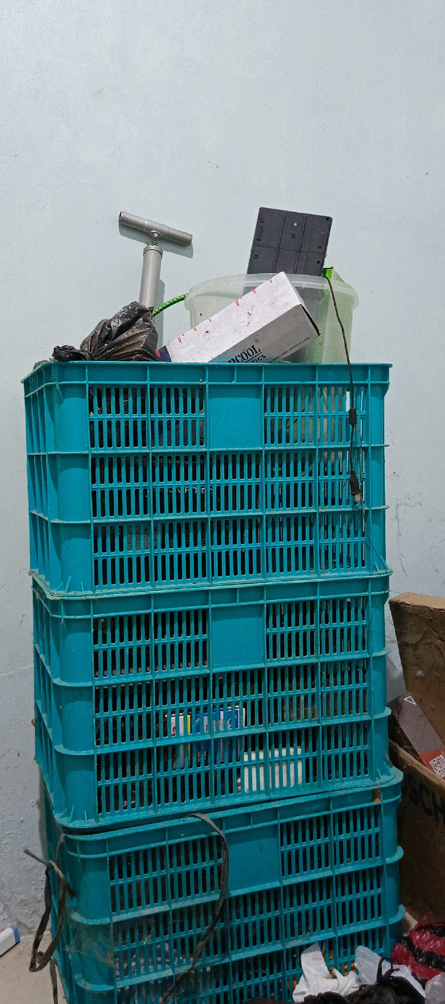 In Pashiebeutong, Indonesia, a stack of three teal plastic crates is positioned against a light blue wall. The crates are filled with various items. The top crate contains a crumpled black plastic bag, a white box with red and black lettering that reads "COOL TICS," a clear plastic container, and a black electronic device with a grid pattern. A silver T-shaped tool with a spring mechanism rests on top of the crates, leaning against the wall. The middle and bottom crates are filled with an assortment of objects, including what appear to be cylindrical items, some with colorful labels, and other indeterminate items. A dark brown cardboard box is partially visible to the right of the crates, and the lower portion of the crates is in contact with the floor, which is out of focus. A dark string is tied around the bottom crate. The lighting appears to be ambient, suggesting an indoor setting.