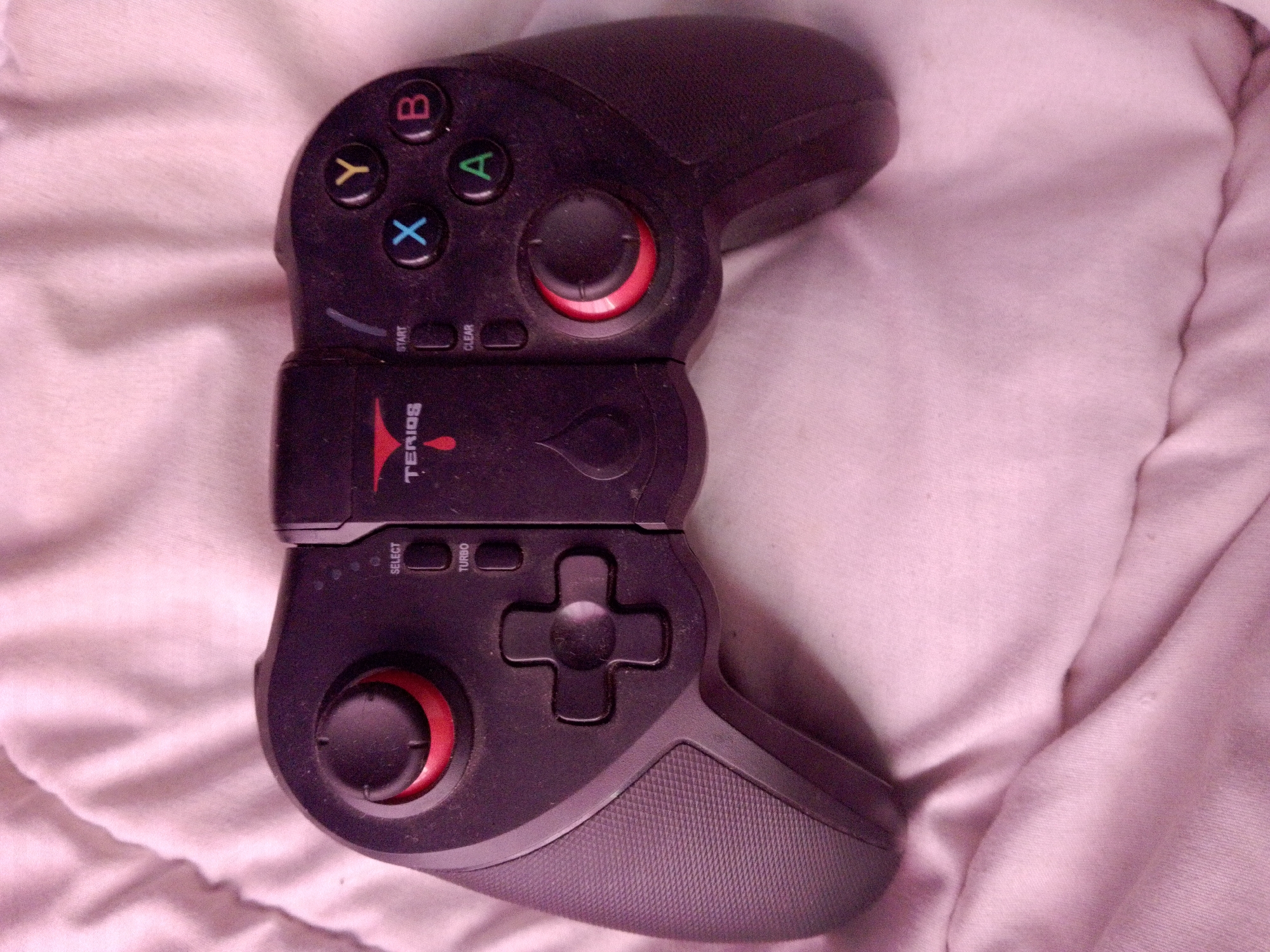 A black game controller rests on a pink blanket. The controller has a directional pad and buttons labeled X, Y, A, and B. It also features two joysticks, start and select buttons, and a turbo button. The "TERIOS" brand name is visible in the center of the controller. The image is a close-up shot and does not provide any information about the location of Inhulo-Kamianka, Ukraine.