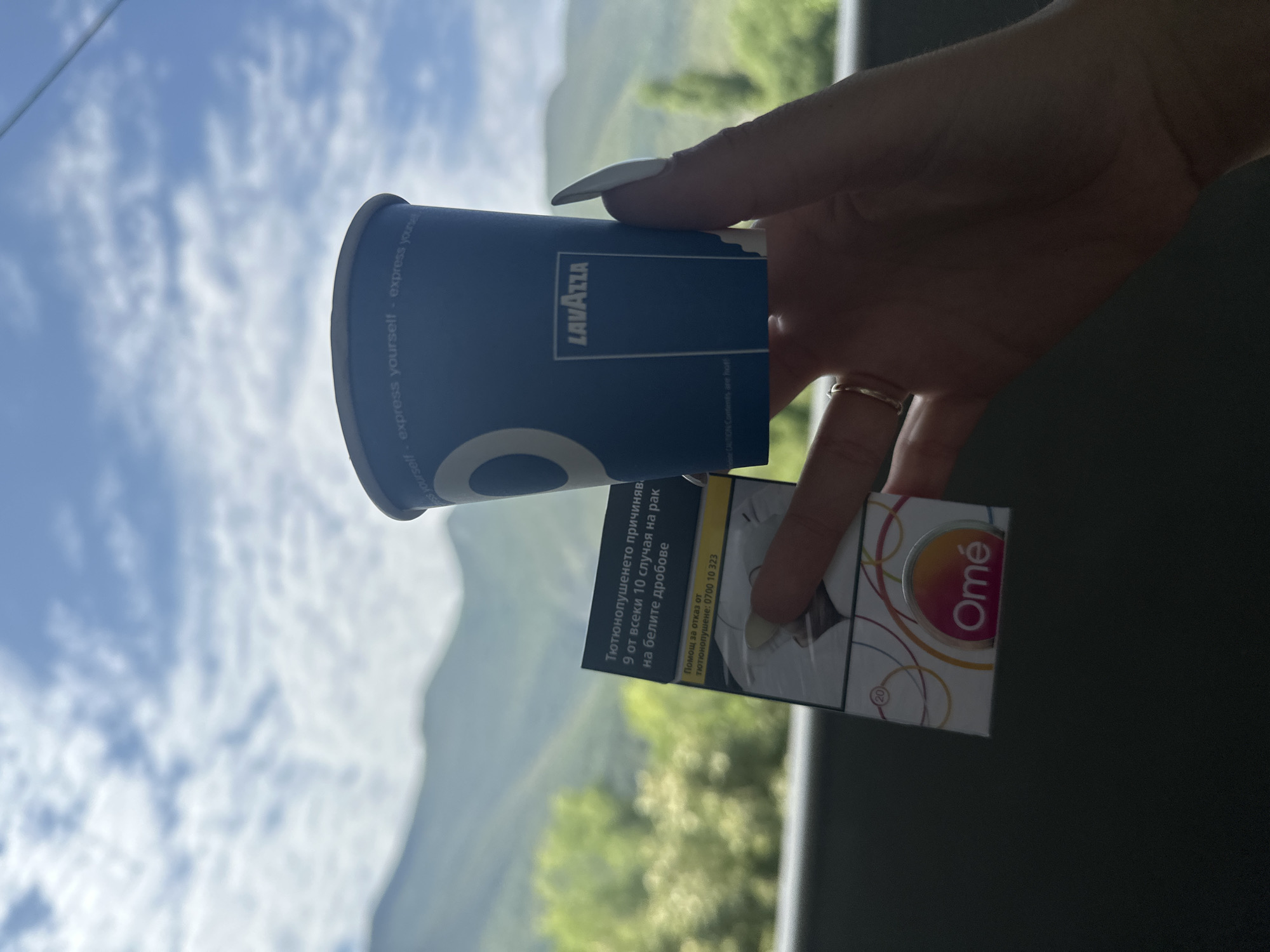 An outdoor, close-up, vertical shot captures a person's hand holding a blue disposable coffee cup with the "Lavazza" logo and a pack of "Omé" cigarettes. The hand has long, manicured nails and a thin gold ring on the index finger. The background features a hazy, green, mountainous landscape under a cloudy, blue sky. The image does not provide any specific geographical markers or elements that would allow for the identification of its location within Vratsa, Bulgaria.