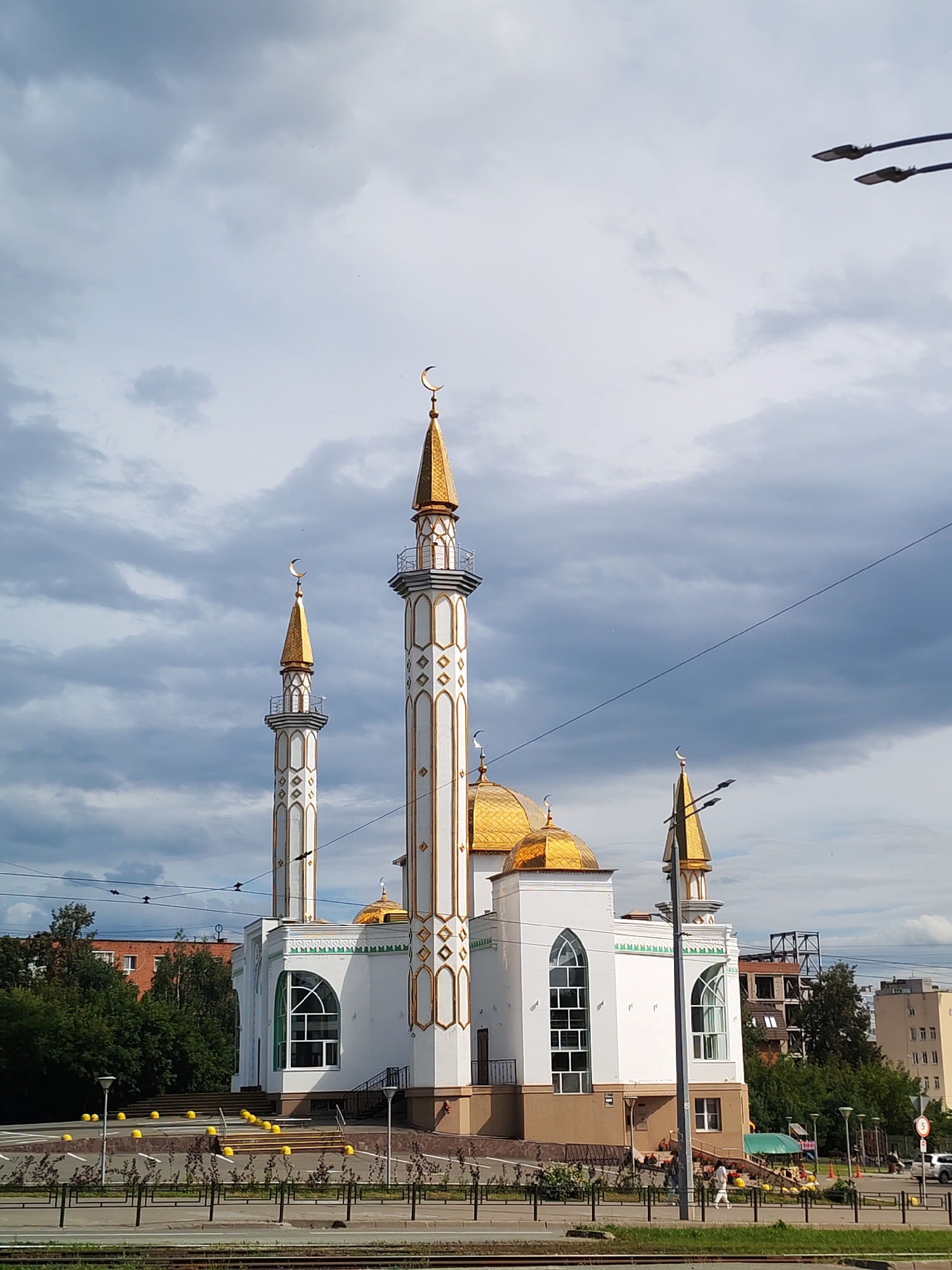 This is a daytime, eye-level, medium shot of the Sobornaya Mosque in Izhevsk, Russia. The mosque is a white building with gold domes and minarets, decorated with intricate geometric patterns. The sky is cloudy with some patches of blue. In the foreground, there is a street with a traffic light and a few cars, as well as a walkway with yellow bollards and some people walking. Trees and other buildings can be seen in the background.
