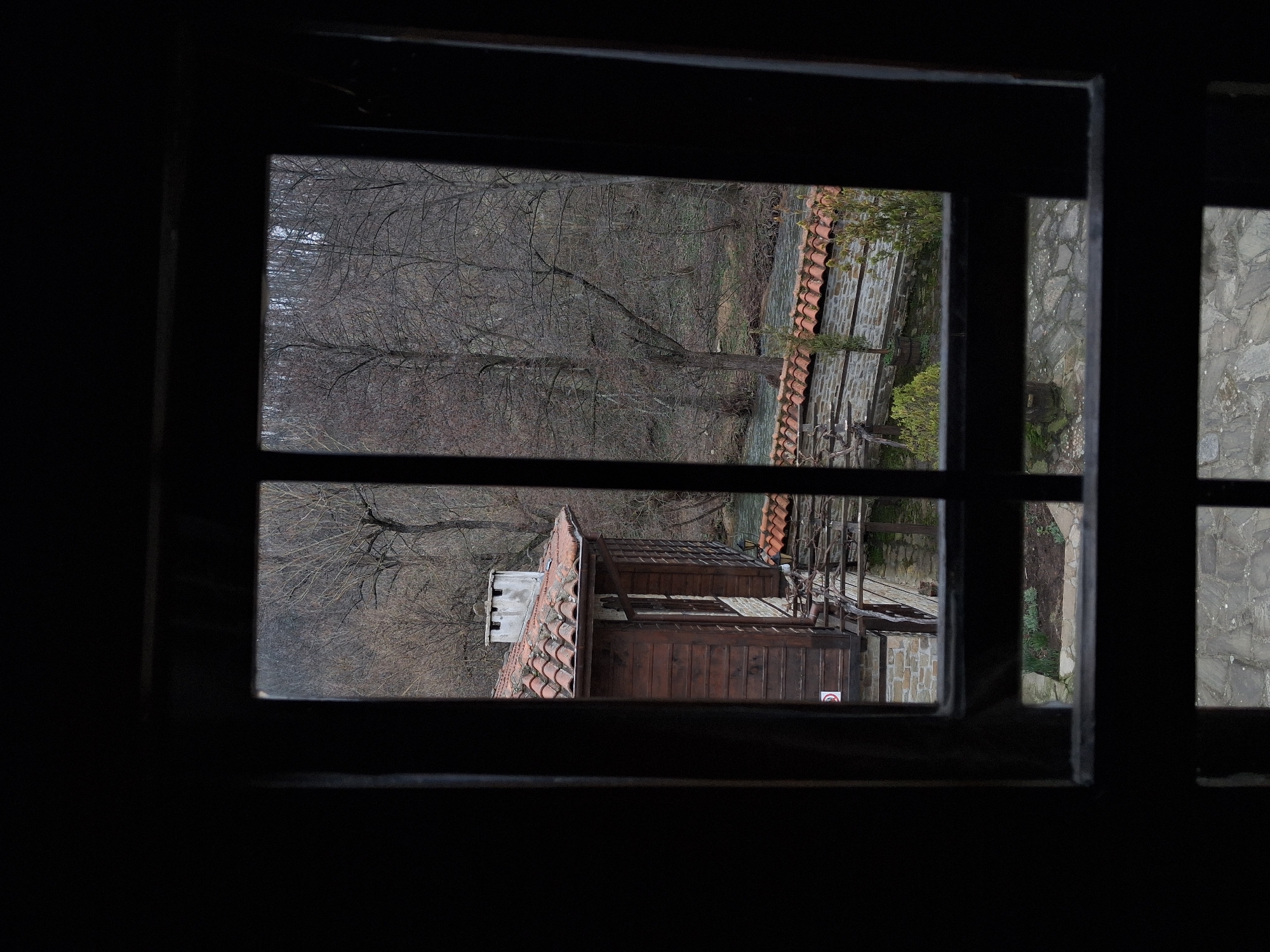 A view from a dark interior through a wooden window frame shows a building with a red tile roof, a forest with bare trees, and a stone wall. The location is Katunishte, Bulgaria.