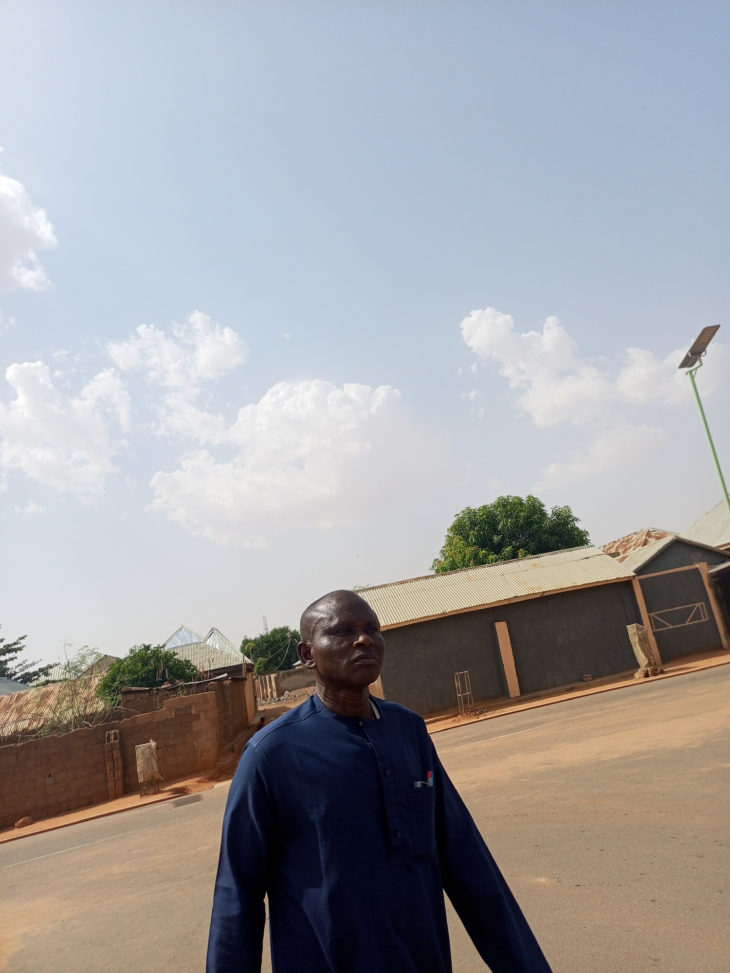 The photograph is an outdoor shot taken in what appears to be a neighborhood in Tempure, Nigeria.
A middle-aged man is standing on an asphalt road, partially obstructing the view of the street. He is wearing a dark blue long-sleeved shirt with a small object or pen in the breast pocket. He is looking off-camera to the right.
Behind the man are various buildings, including a long, dark grey structure with a corrugated metal roof. The surrounding environment includes trees, brick walls, and a tall green lamppost with a solar panel. The sky is light blue with scattered white clouds. The ground, including the road and surrounding area, has a reddish hue, likely due to the soil type in the region. The composition is angled, suggesting that the photographer was standing on the side of the road or in motion while capturing the shot.