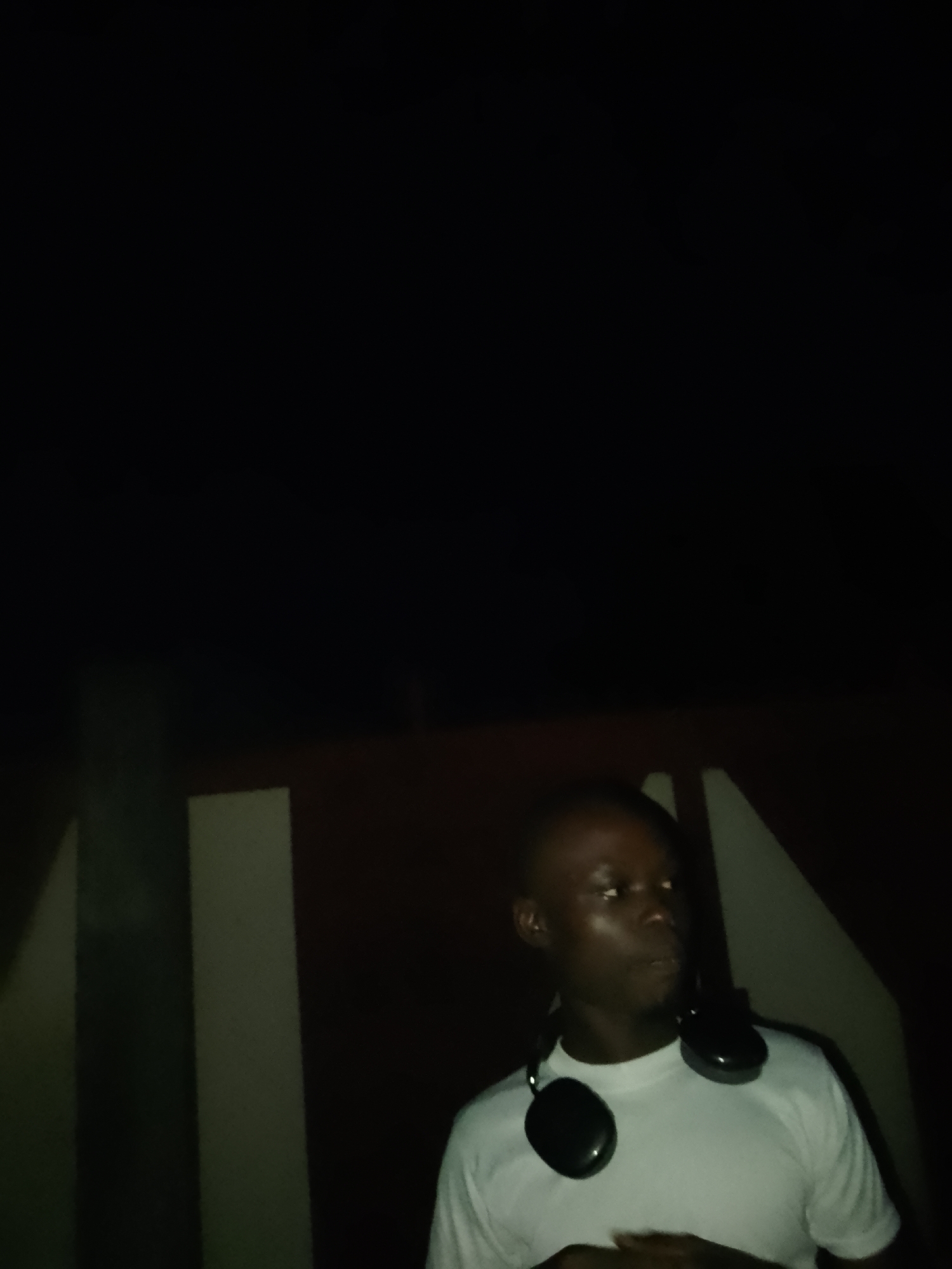 A man wearing a white t-shirt and headphones around his neck stands in front of a dark background with vertical light-colored stripes. His head is turned to the right, and he appears to be looking away from the camera. The lighting is dim, casting shadows on his face. The man is the sole subject in the image. The location is identified as Serti, Nigeria.