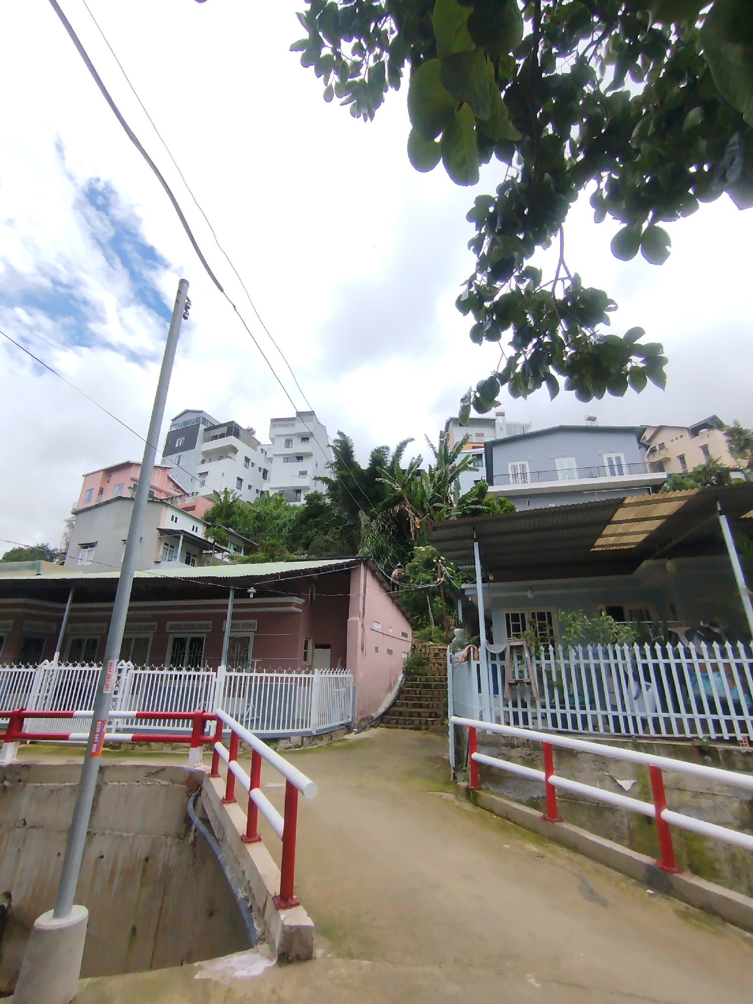 The image captures a residential street or pathway winding uphill through a densely built area on a hillside, characteristic of Da Lat, Vietnam. The time of day is daytime, with an overcast sky showing some patches of blue.
In the foreground, a concrete path leads slightly upward. On the left, a concrete barrier or opening, possibly for drainage, is protected by red and white metal handrails. A grey utility pole stands prominently on the left side of the path, bearing a white sticker with partially visible text, including the phone number "0930 091 087." On the right, more red and white metal handrails line the path, bordering a raised section.
The midground features two main houses: one on the left, painted light pink or peach, and another on the right, light blue or grey. Both homes are single-story with corrugated metal roofs and white picket fences. A set of concrete stairs ascends the hillside to the right of the blue house. Lush green vegetation, including what appears to be banana plants, grows between and around the houses.
Further up the hillside, in the background, several multi-story buildings are visible, constructed on the steep terrain. These buildings vary in color, featuring white, grey, and light pink facades, and many include balconies. Overhead power lines stretch across the scene. A tree branch with dark green leaves hangs into the frame from the top right. No people are visible in the image.