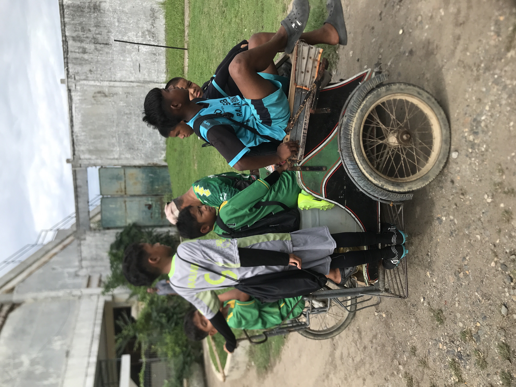 In Lhokseumawe, Indonesia, four young men are seen on a modified bicycle. The bicycle has a sidecar-like attachment. One boy is riding, two are passengers in the sidecar, and the last is in the cart attached to the bike. The location appears to be an outdoor area, possibly near a building or structure. The background includes greenery, and the ground is unpaved. The day appears to be overcast. The clothing worn by the boys suggests they are likely students in a relaxed setting.