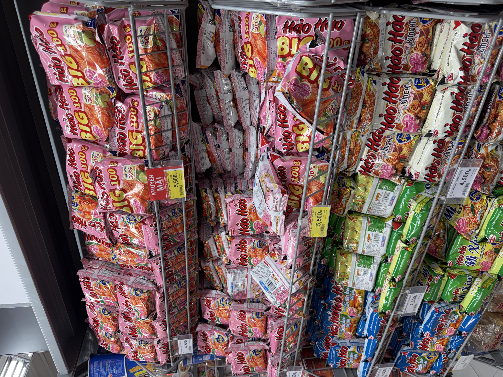 The image shows a retail shelf displaying various packages of instant noodles in a supermarket. The shelves are filled with brightly colored packaging, predominantly pink, yellow, and blue. The products are neatly arranged in rows. The brands visible include "Hao Hao" and "Big Hao Hao". Some packages have promotional labels indicating discounts or special offers. In the foreground, price tags are visible, showing prices such as "4,500đ" and "5,500đ". The lighting suggests an indoor environment with fluorescent overhead lights typical of a supermarket. No people are visible in the image. The scene appears to be a standard retail display of consumer goods.